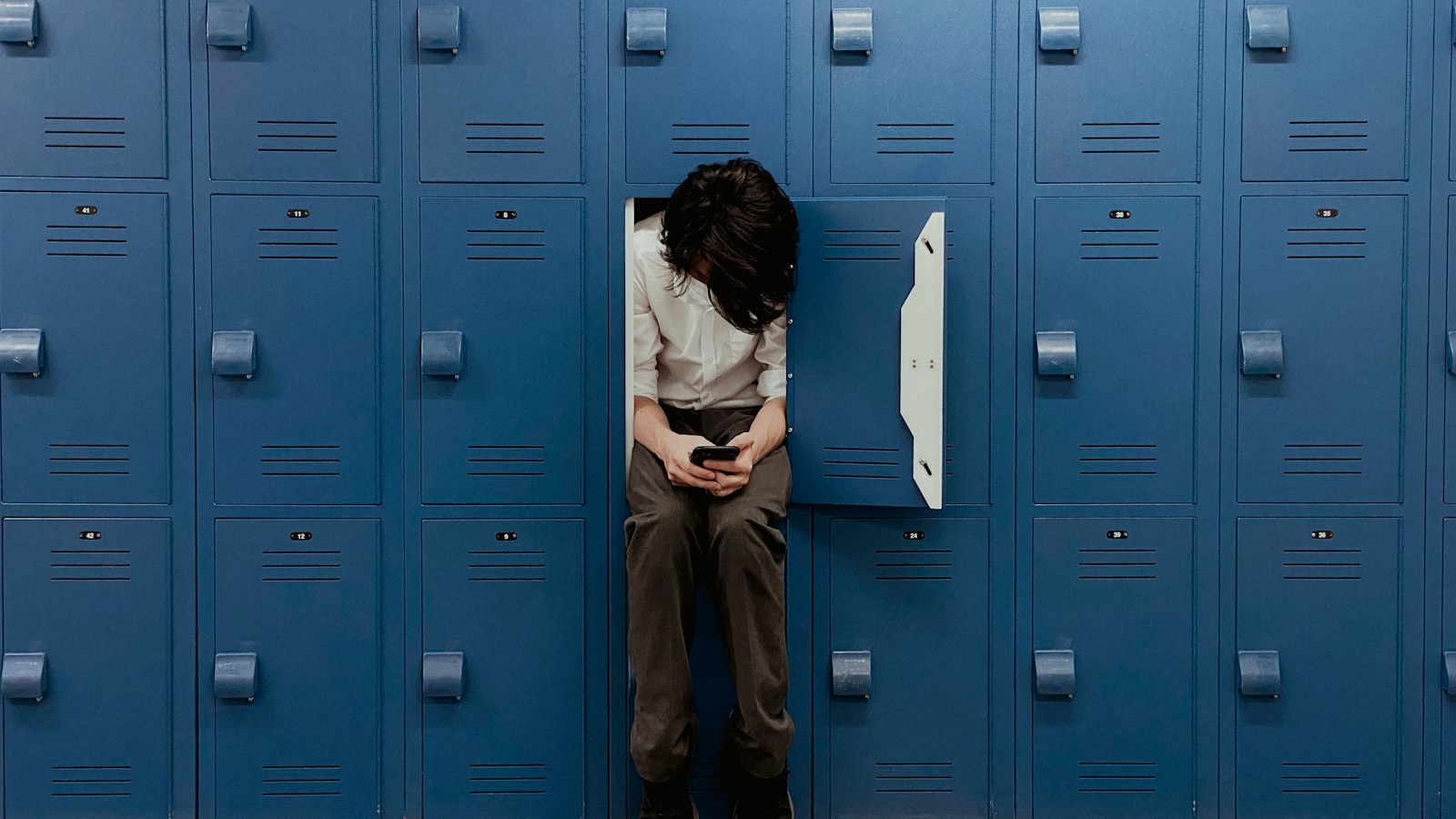 A person in a white shirt sits folded inside an open blue locker in a wall of numbered lockers, looking at a phone