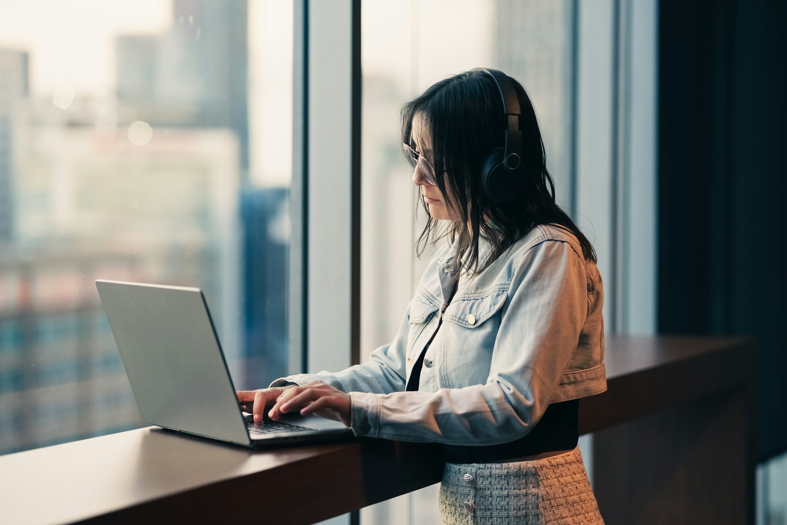 A woman in headphones and a denim jacket types on a laptop at a window counter above a city
