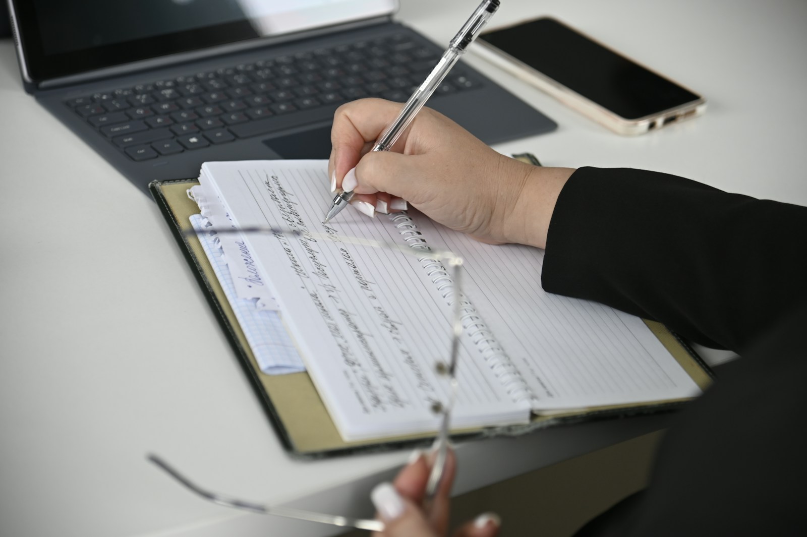 A hand writes notes with a clear pen in a spiral notebook, next to a laptop keyboard and a phone