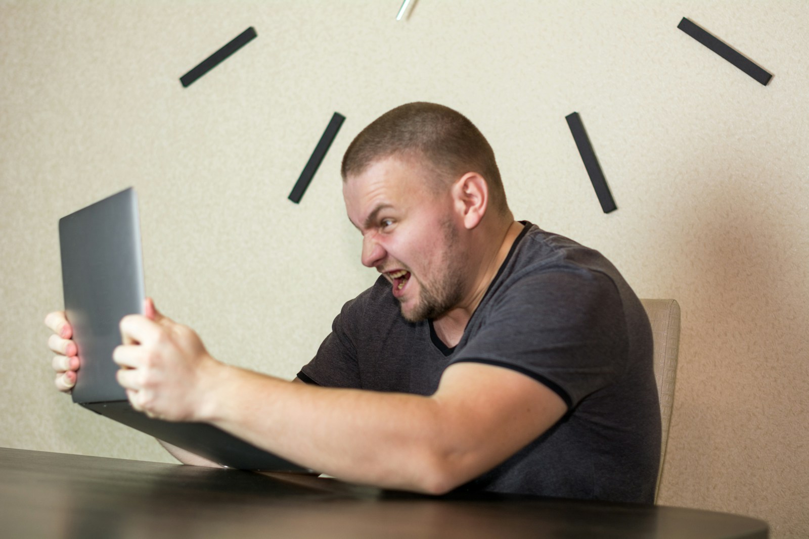 A man in a dark T-shirt grips a laptop with both hands and shouts at its screen