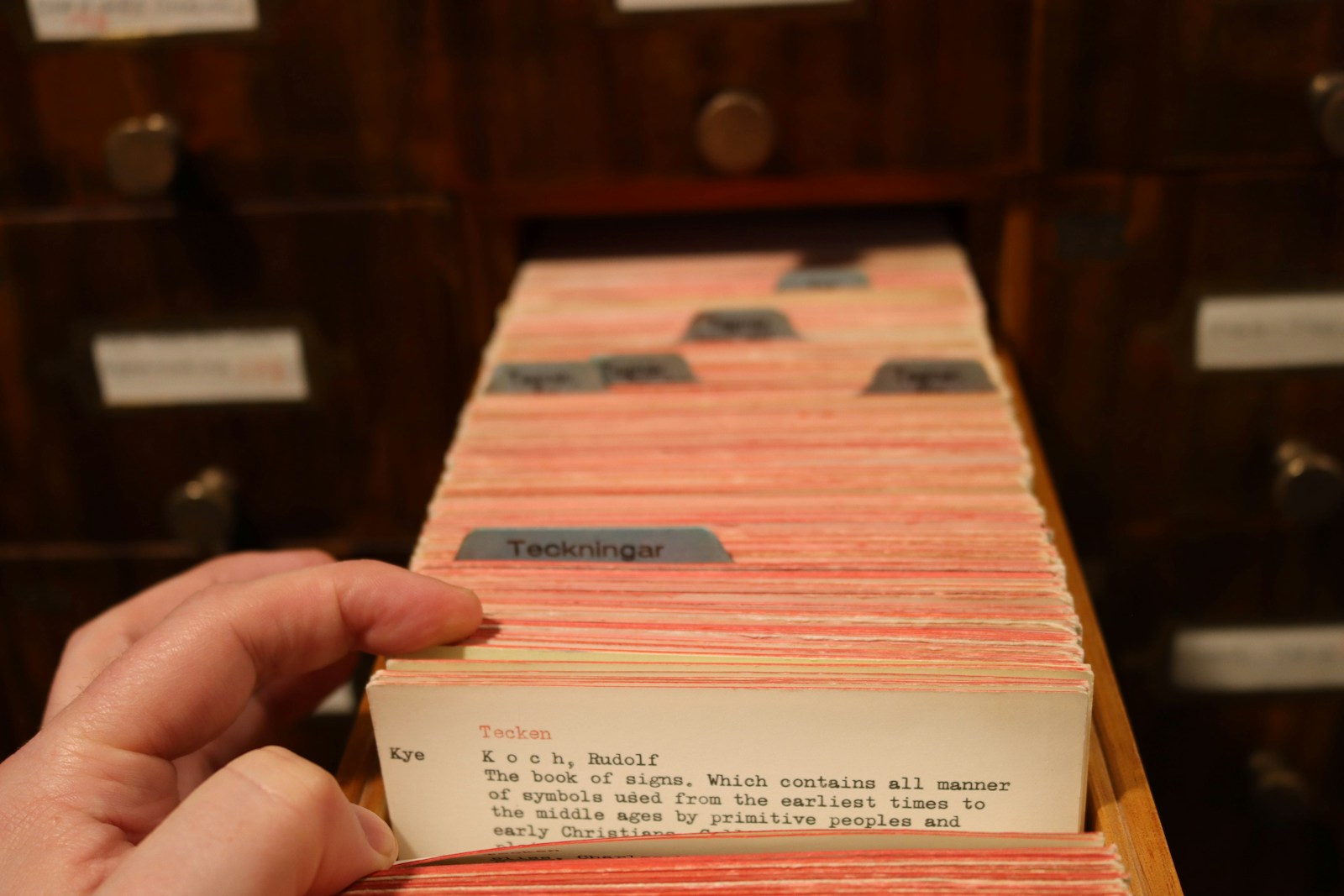 A hand flips through a drawer of typed library catalogue cards with blue index tabs in a dark wooden cabinet