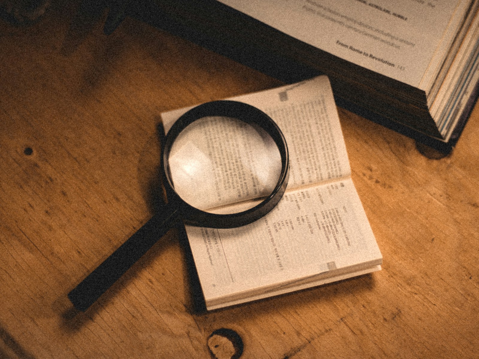 A black magnifying glass rests on a small open book on a wooden table beside a larger open book