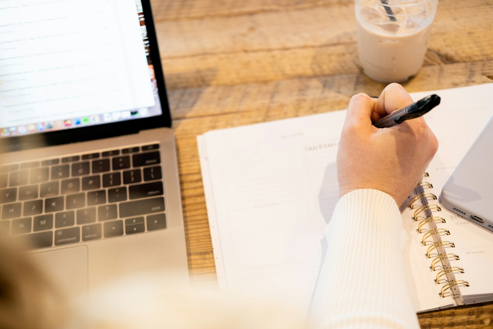 A hand writes in a spiral planner next to an open laptop and an iced drink on a wooden table