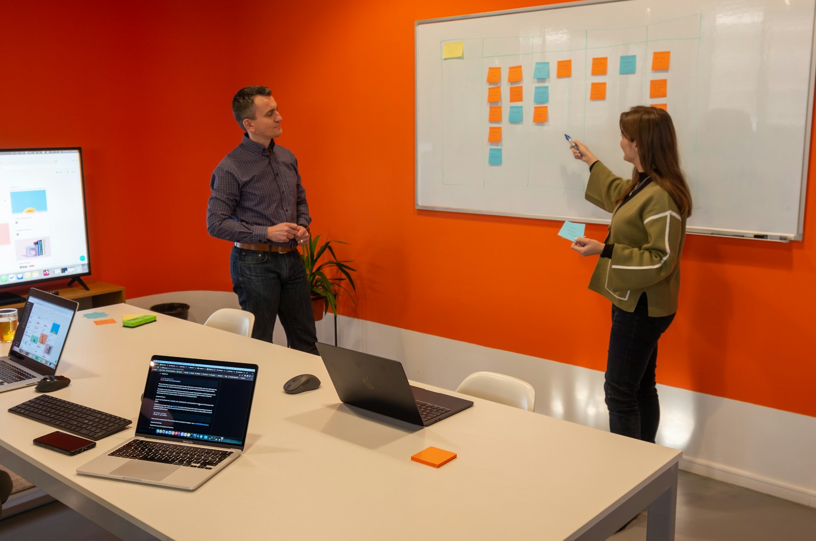 A woman points a pen at a whiteboard covered in orange and blue sticky notes while a colleague watches