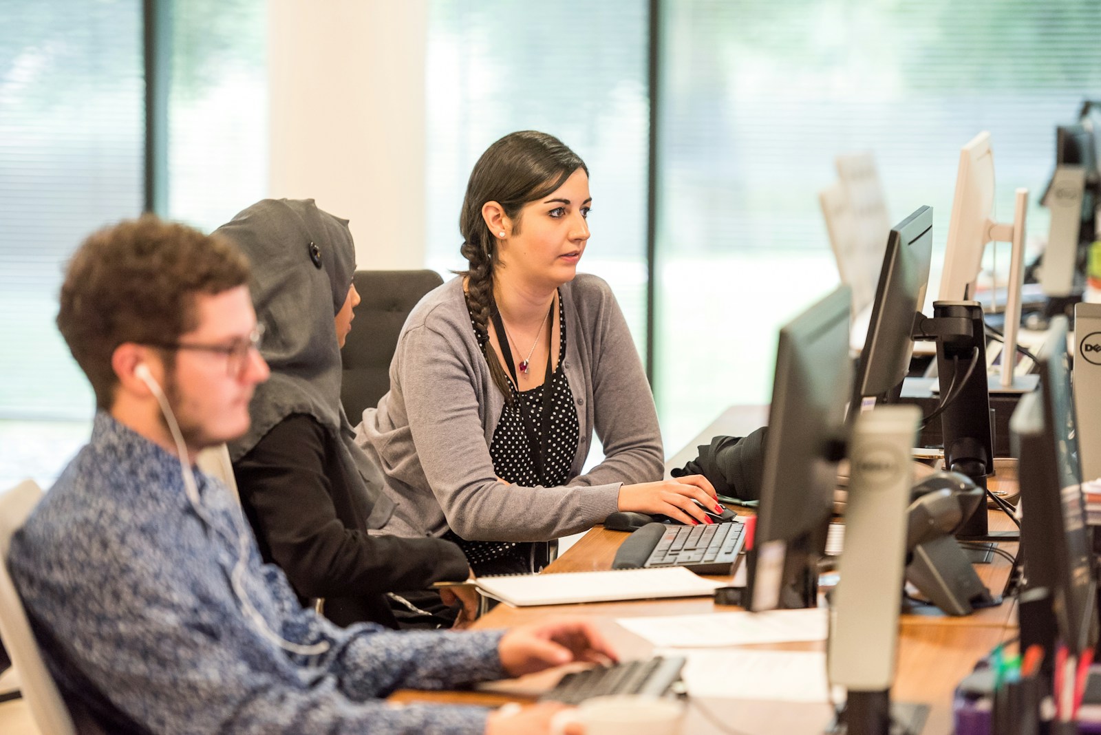 Three colleagues work at a long row of desks with monitors and desk phones in a bright office