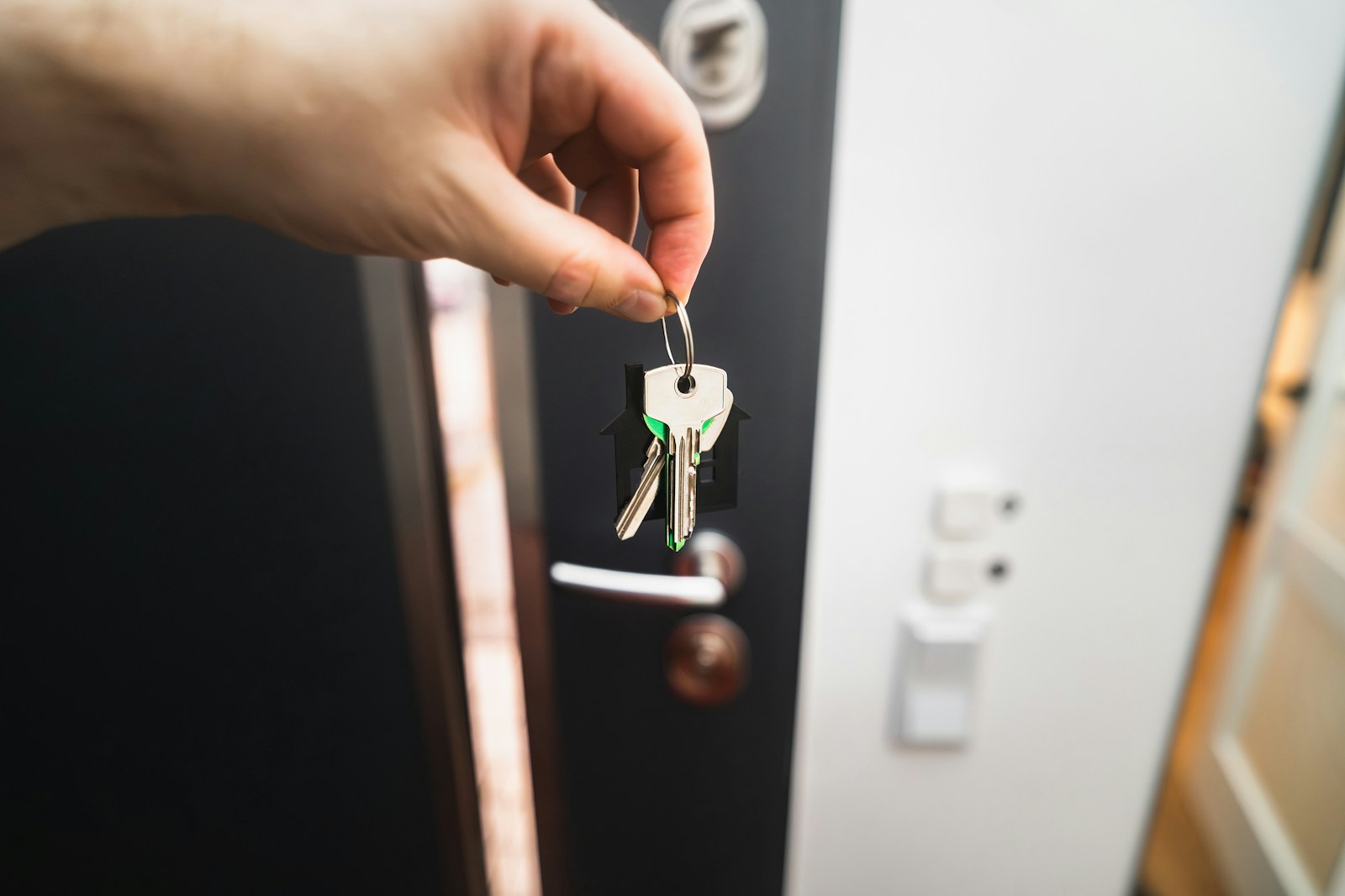 A hand holds up a ring of keys in front of a dark front door with a light switch beside it