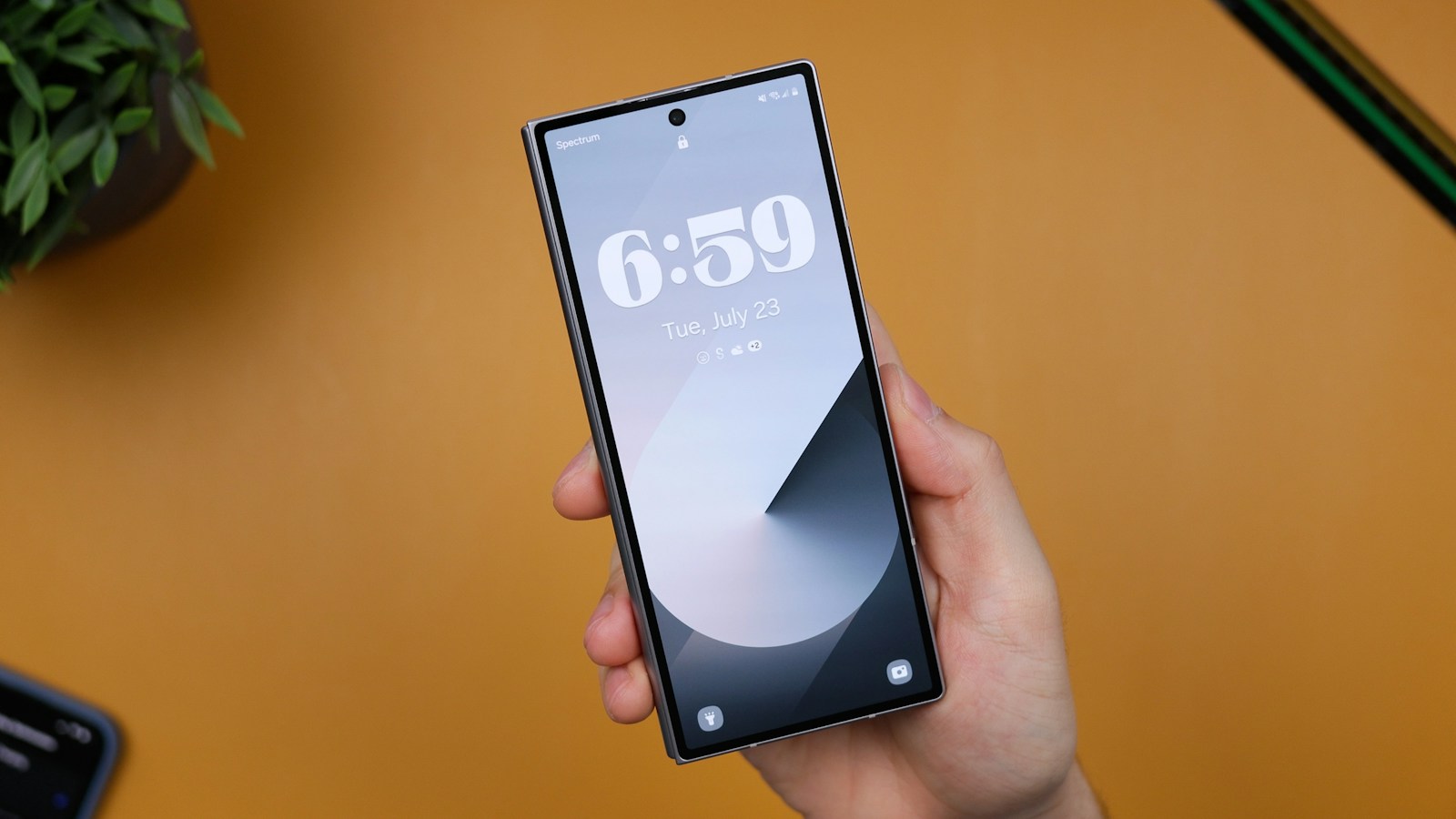 A hand holds a smartphone showing its lock screen above an orange desk with a plant in the corner
