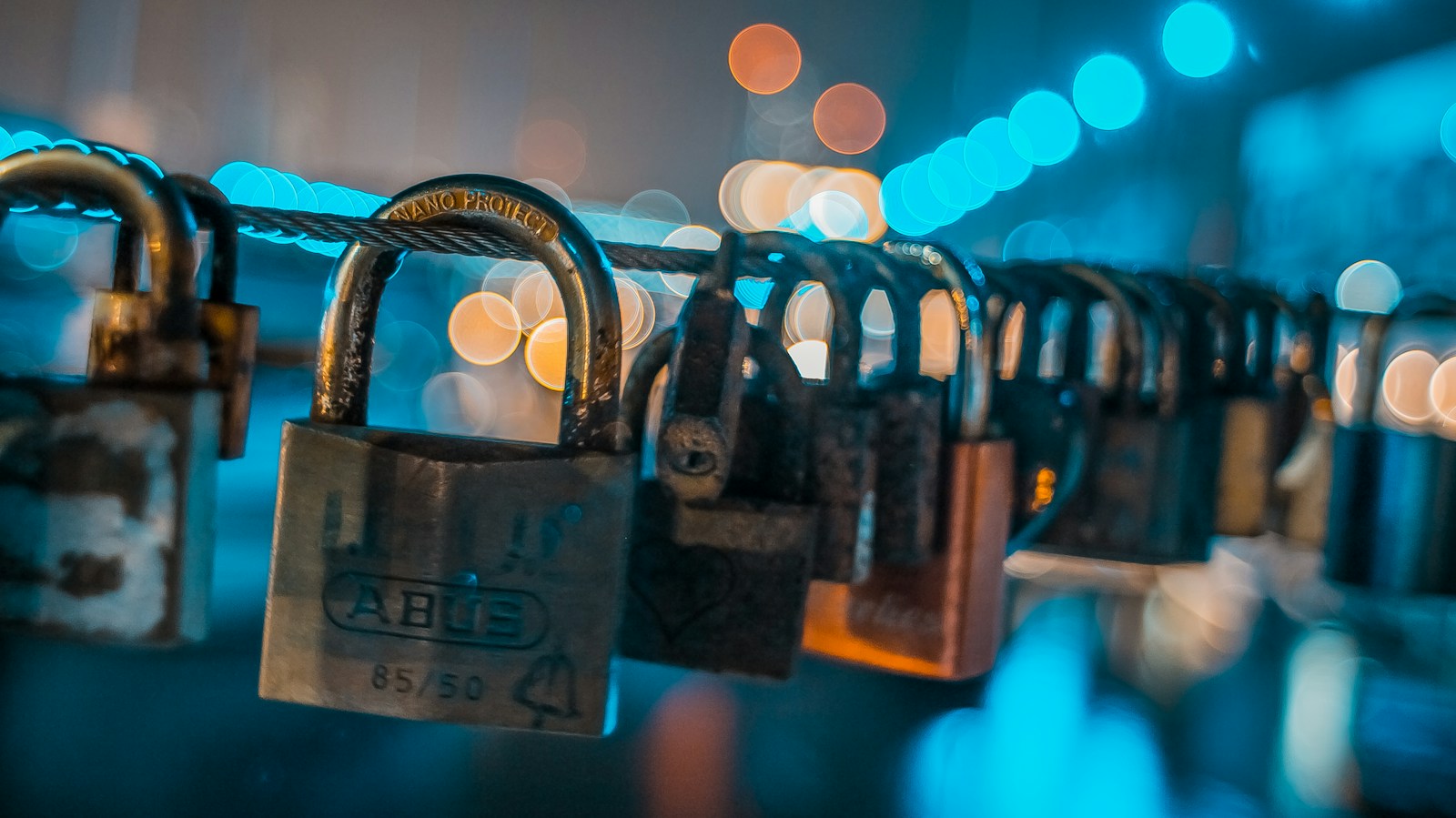 A row of padlocks locked onto a steel cable at night, with blurred orange and blue city lights behind them