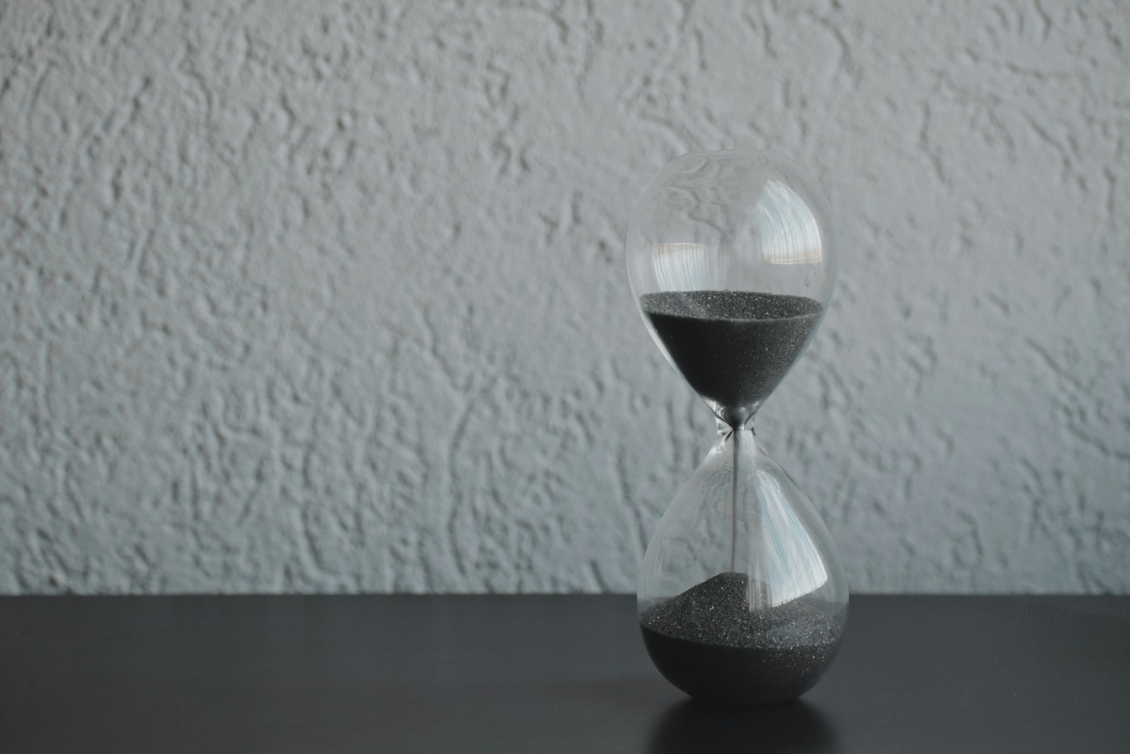 A glass hourglass with dark grey sand running through it, standing on a dark table against a white wall