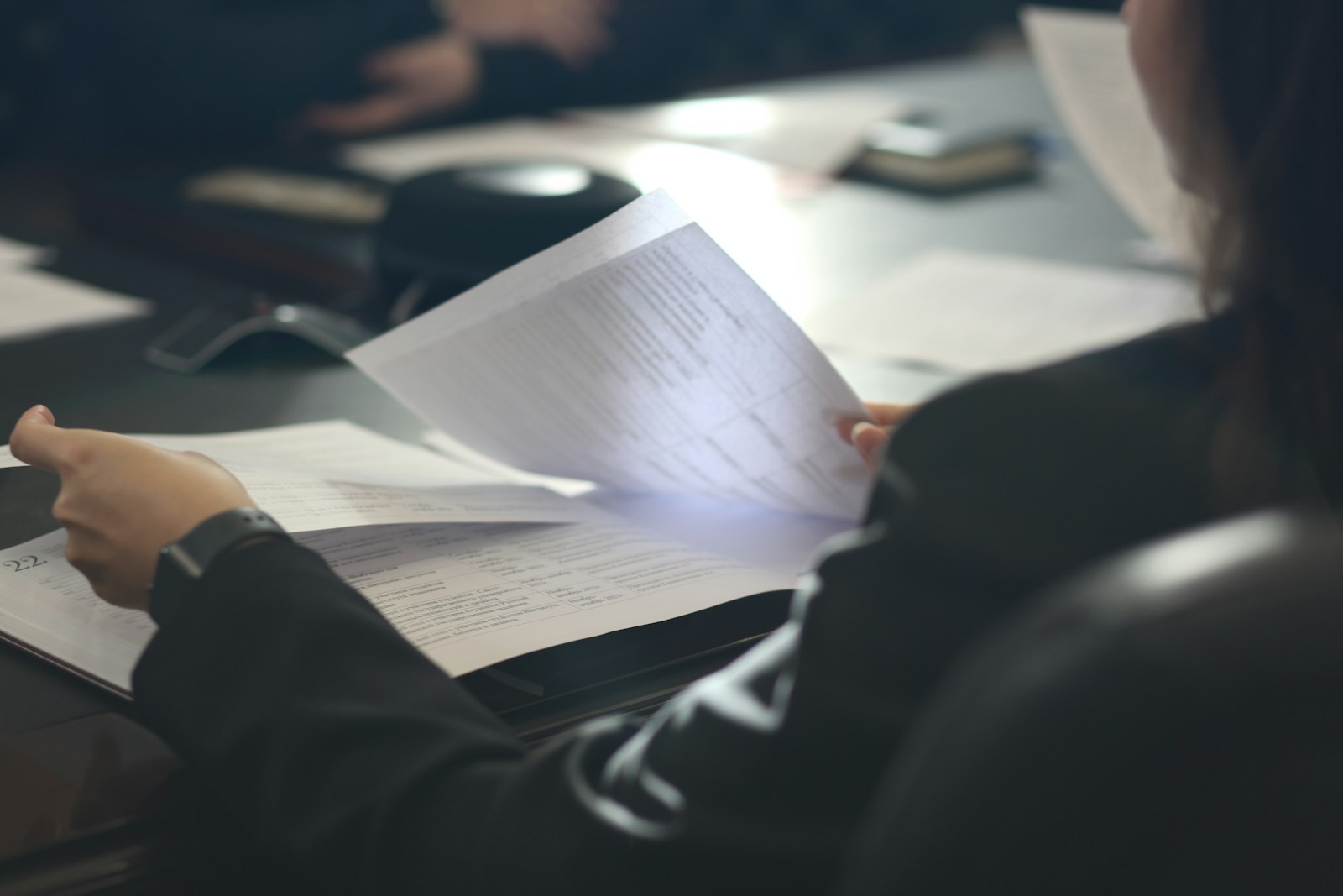 A person in a dark top reads through printed pages of tables and text at a meeting table