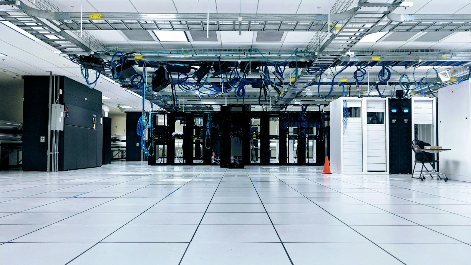 A data center floor with white raised tiles, rows of black server racks and blue network cables hanging overhead