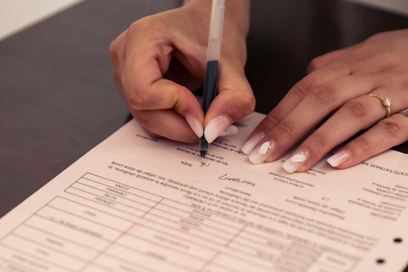 A woman's hands sign a printed form with a ballpoint pen on a dark wooden desk