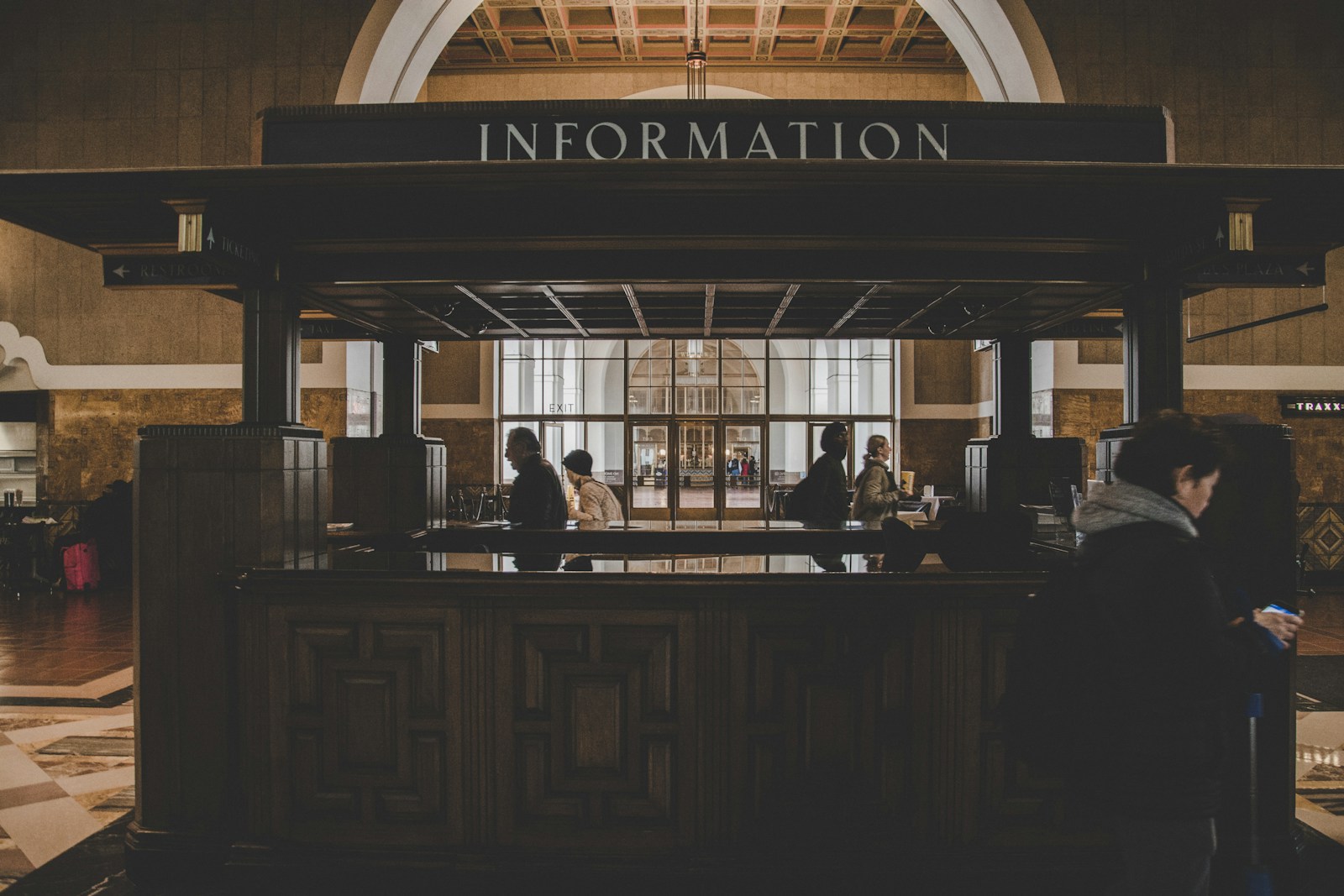 An old wooden kiosk under a large Information sign in a dim, high-ceilinged hall, with people at its counter