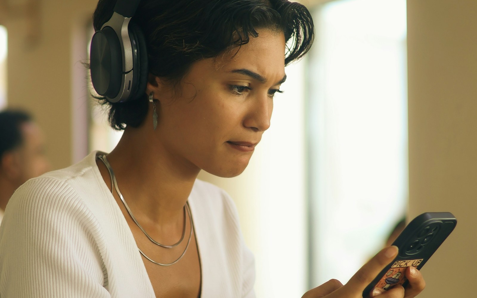 A young woman in headphones and a white cardigan frowns at her phone, puzzled by what she is reading