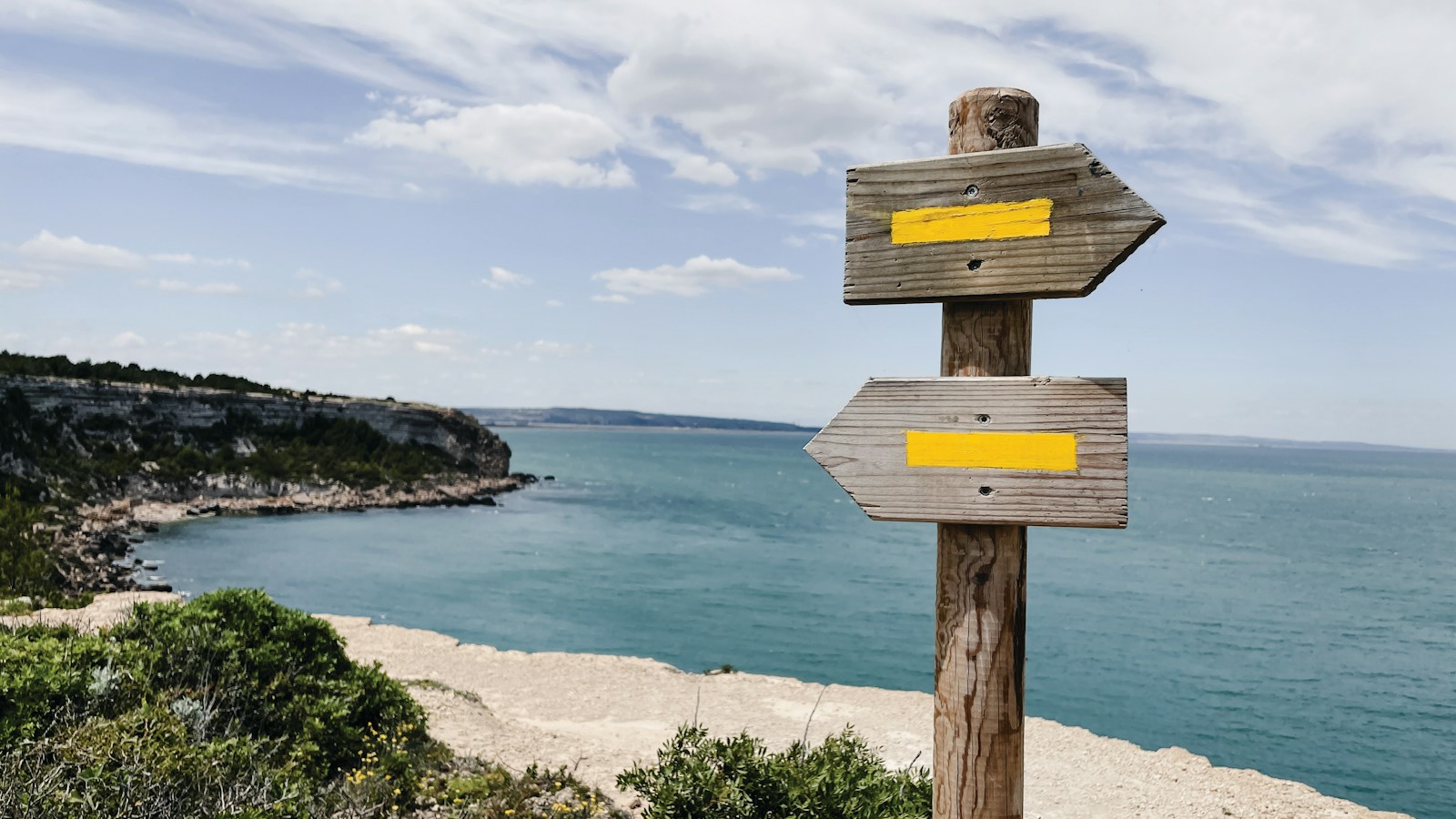 A weathered wooden signpost with two arrows pointing opposite ways stands on a clifftop above a turquoise sea