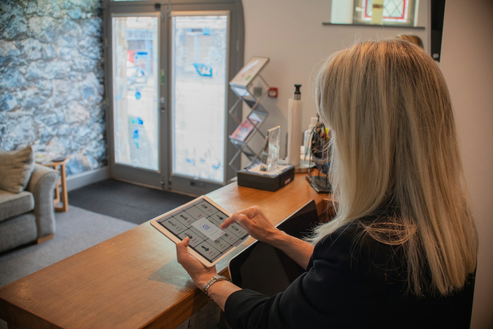 A woman taps a tablet at a wooden reception desk beside a leaflet stand, with a waiting area behind her