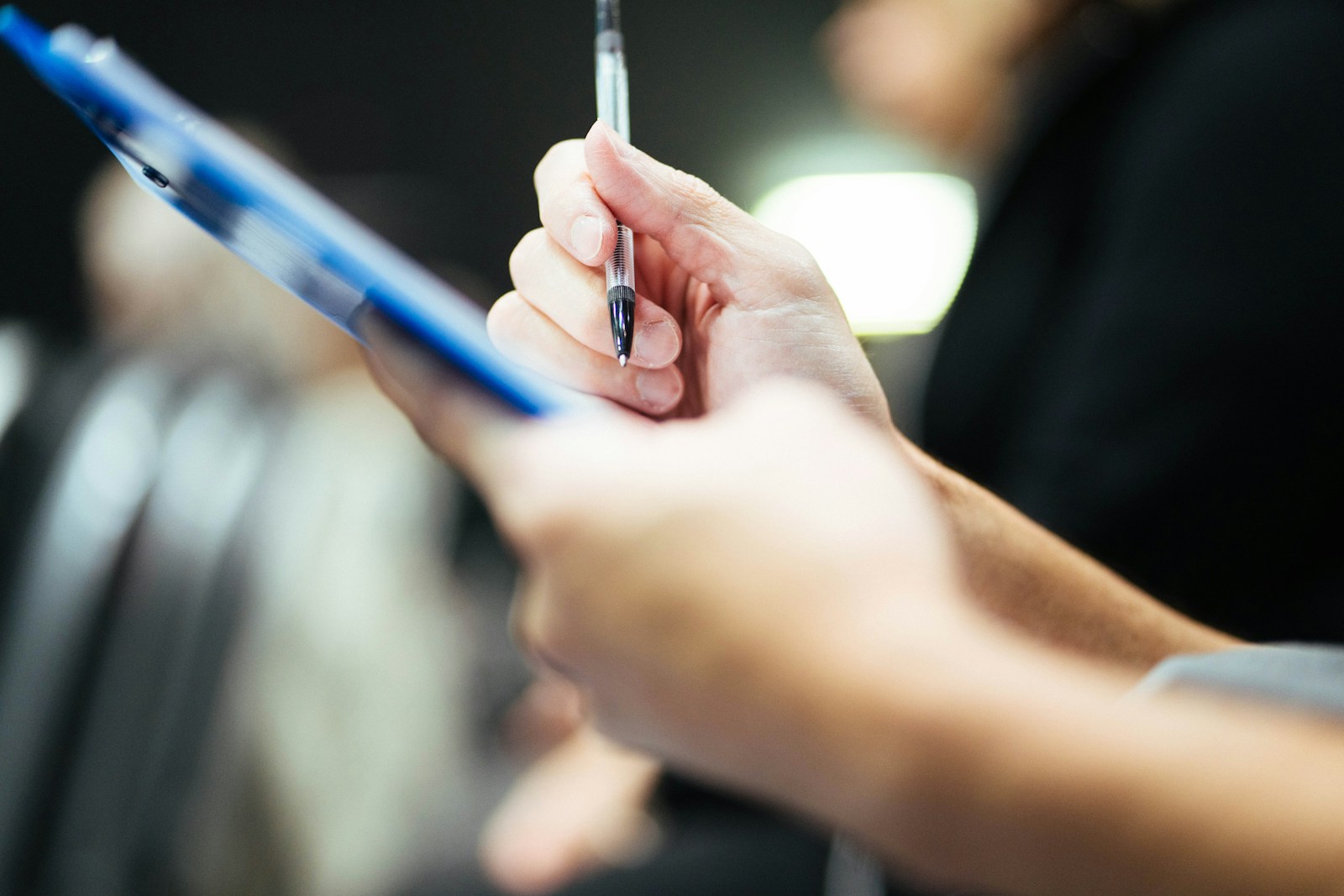 Close-up of a hand holding a ballpoint pen above a blue clipboard, ready to fill in a paper form