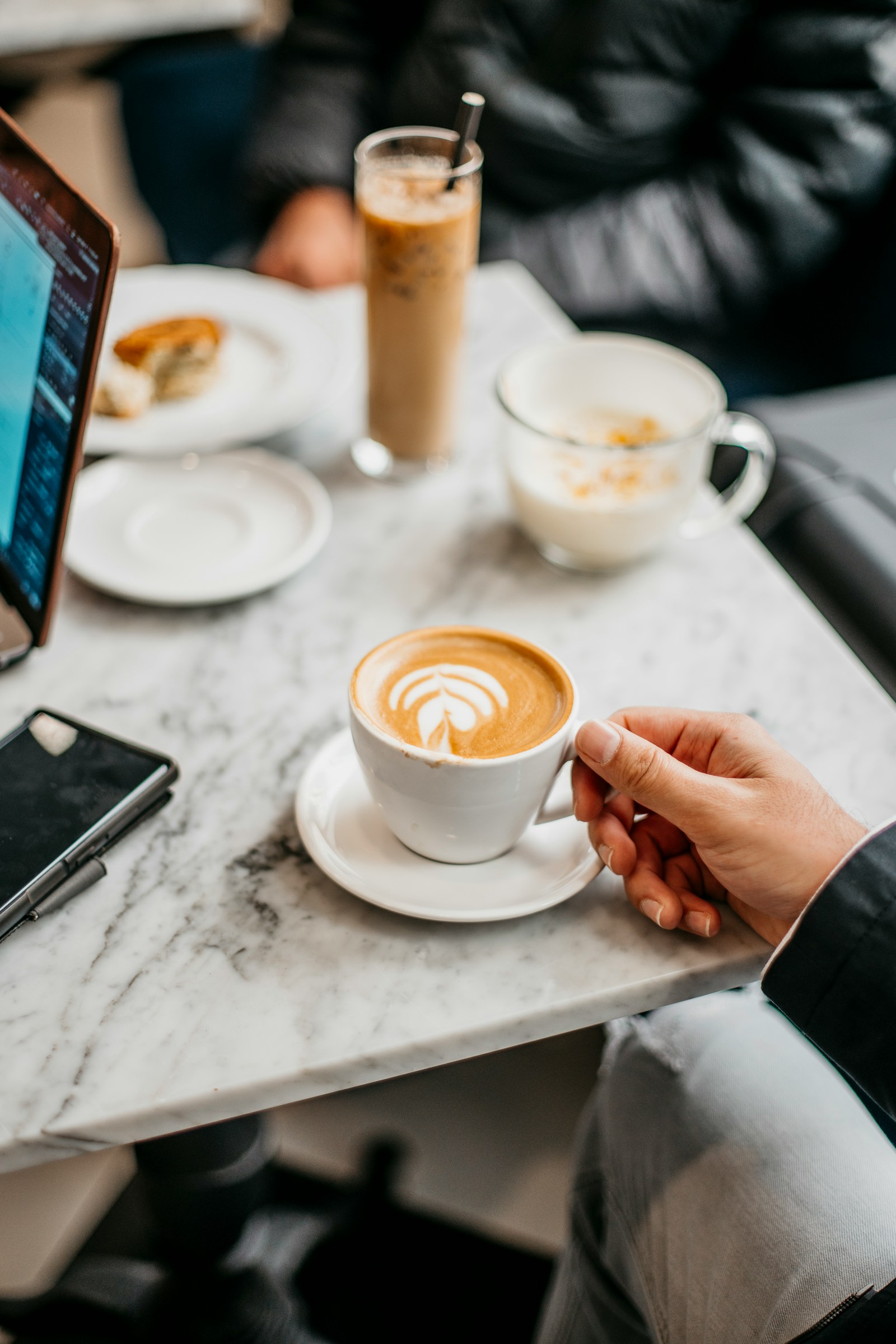 A hand holds a latte with leaf foam art on a marble café table beside a phone and an open laptop