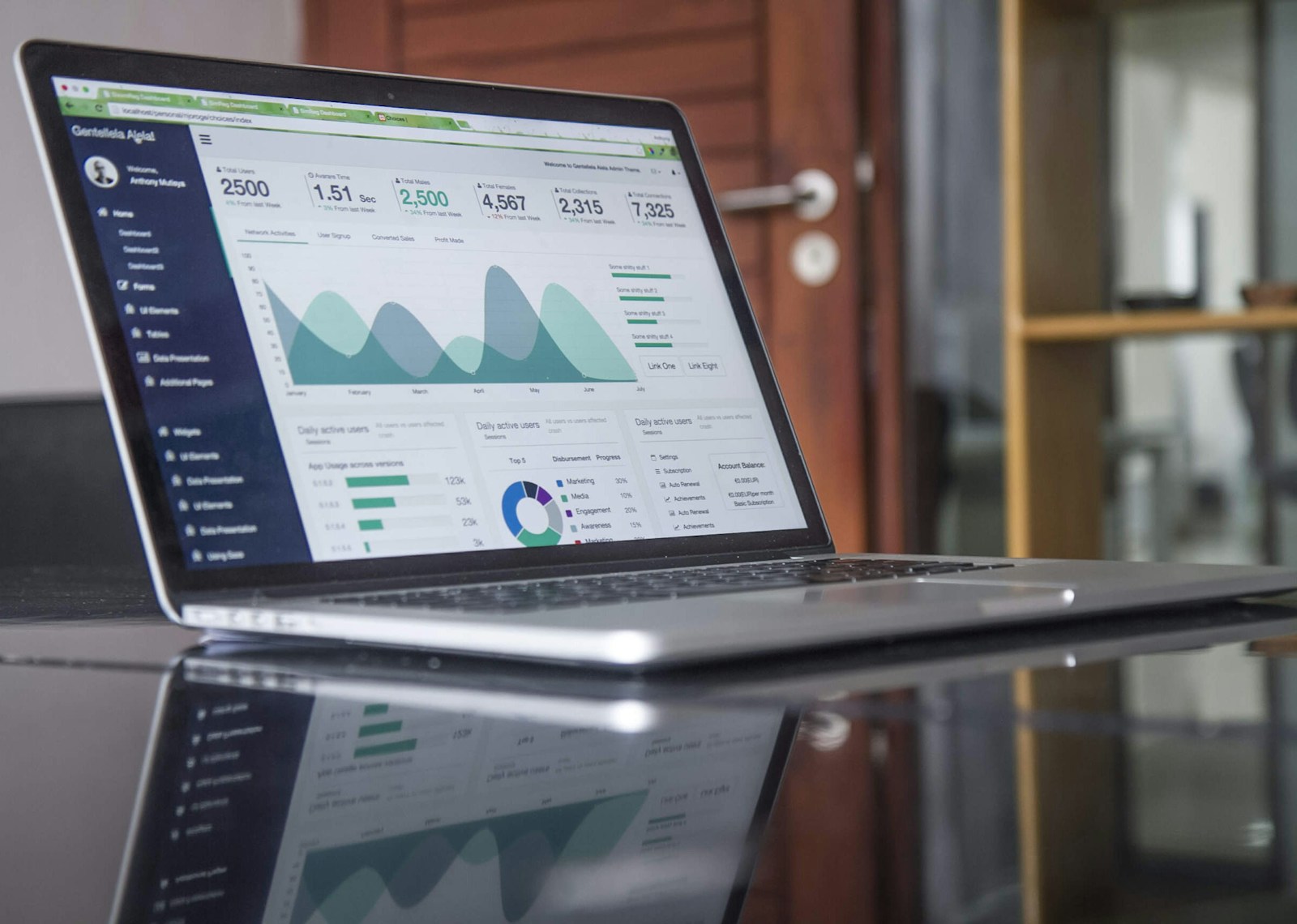 An open laptop on a glass table displays an admin dashboard with user totals, area charts and a donut chart