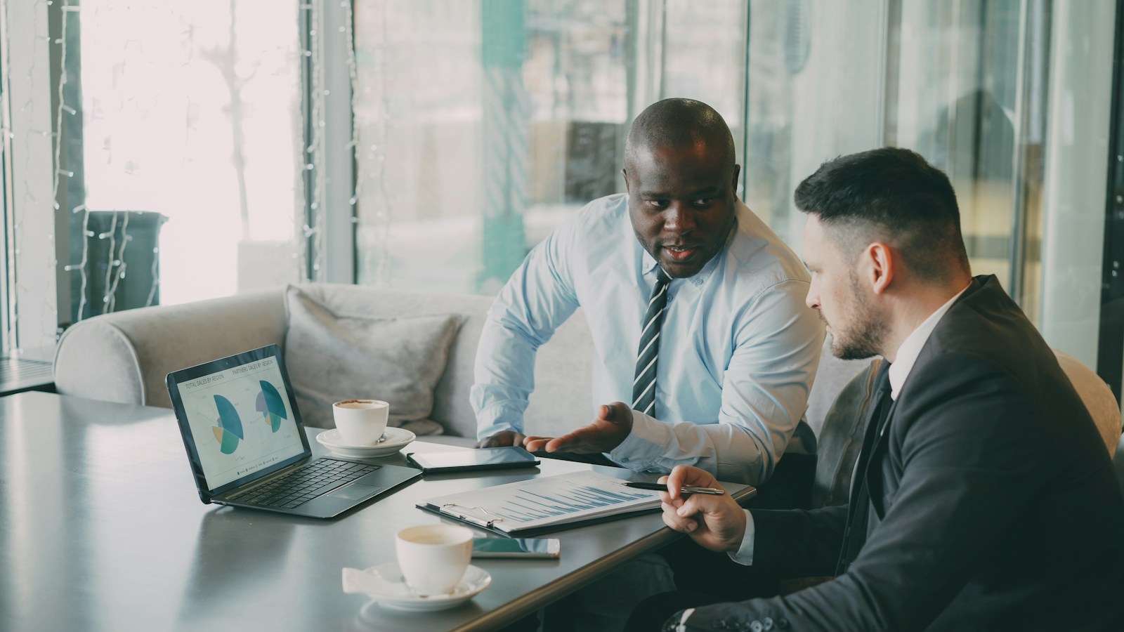 Two men in business clothes discuss sales charts on a laptop and a clipboard at a cafe table with coffee cups