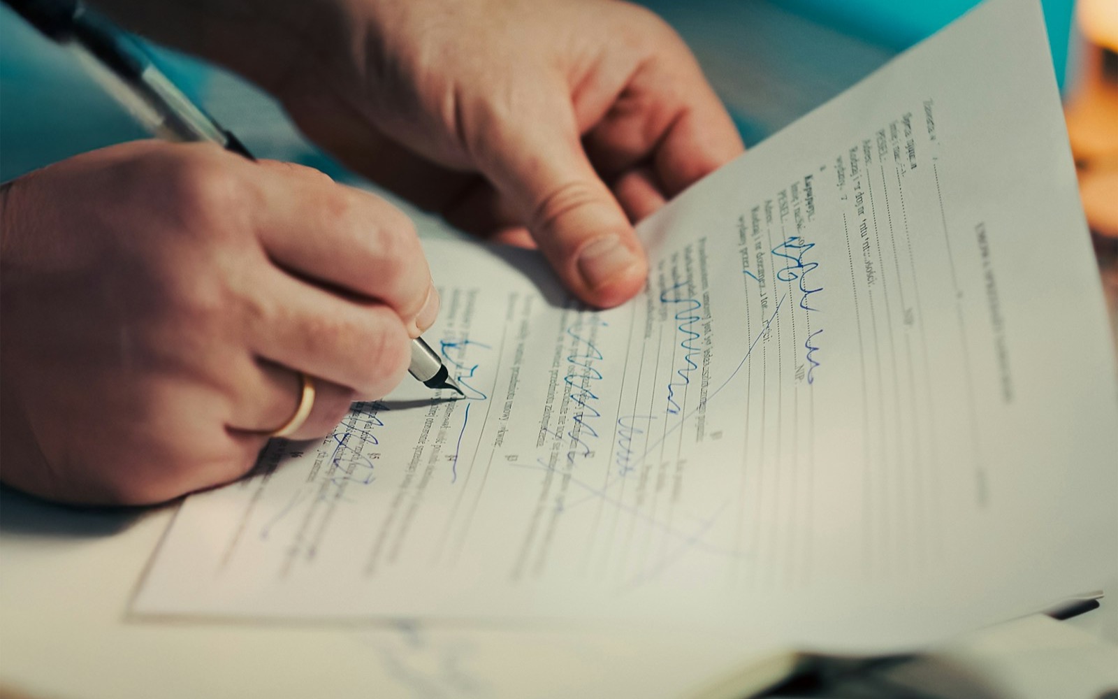 Close-up of two hands, one wearing a gold ring, signing a printed form with a fountain pen in blue ink
