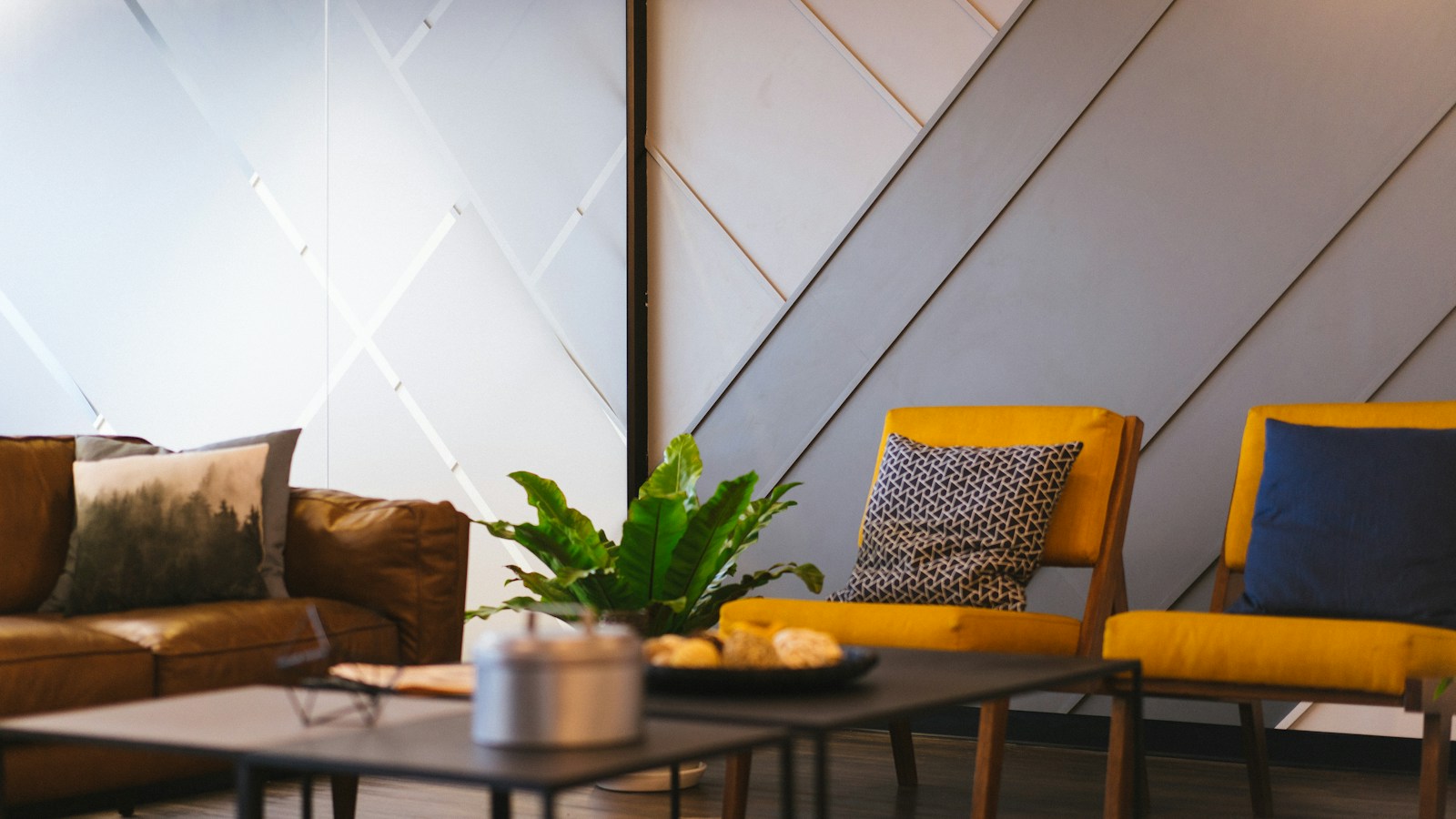 Two empty yellow armchairs and a brown leather sofa around low tables in a quiet lounge with a potted plant