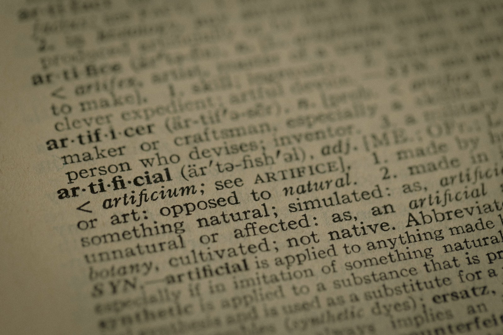 A close-up of a printed dictionary page with the entry for the word artificial in sharp focus