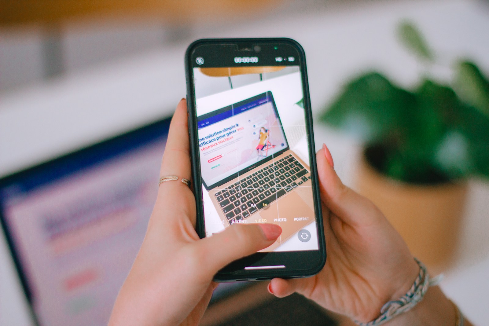 Two hands hold a phone in camera mode that frames a laptop showing a website, with a potted plant behind