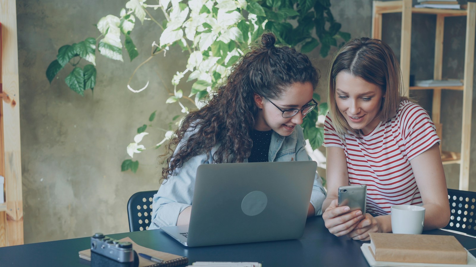 Two women at an office table look at a phone together, with an open laptop in front of them