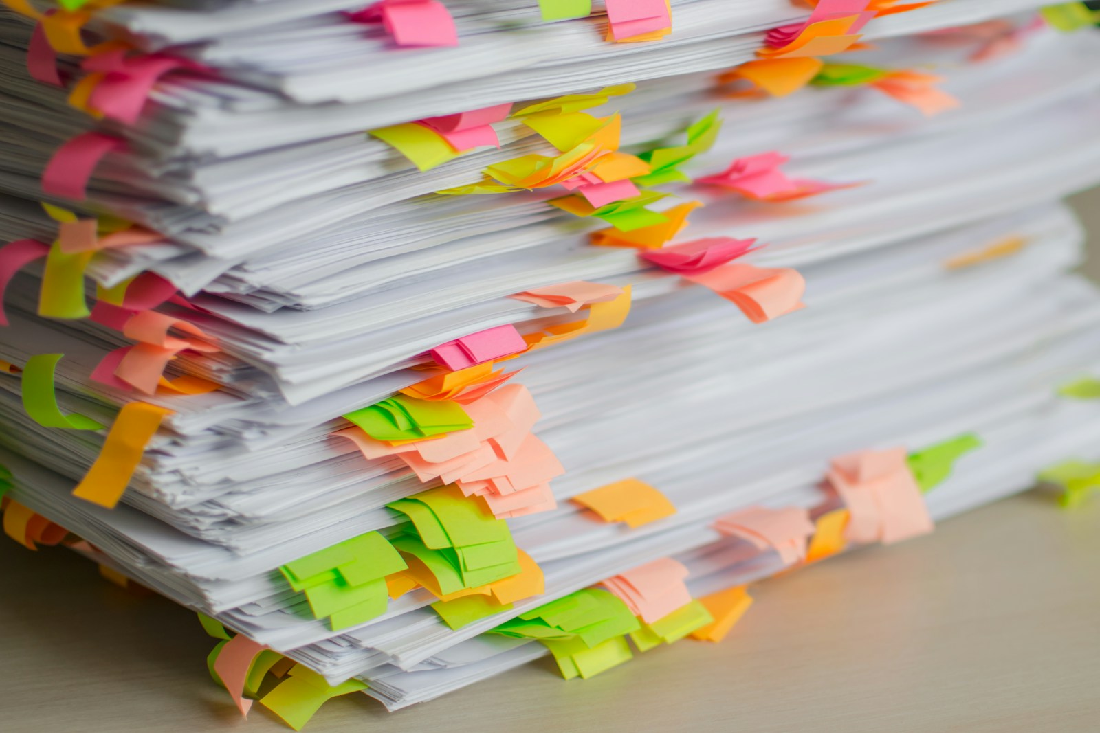 A tall stack of printed papers on a desk, flagged with pink, orange, yellow and green sticky tabs