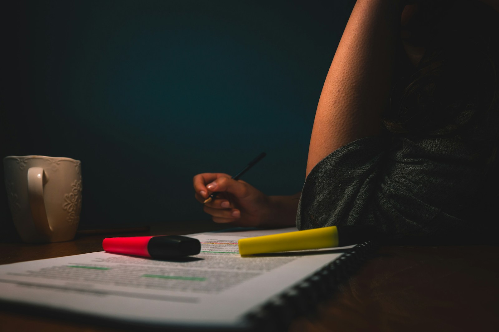 A person with a pen reviews a highlighted printout beside pink and yellow highlighters and a mug