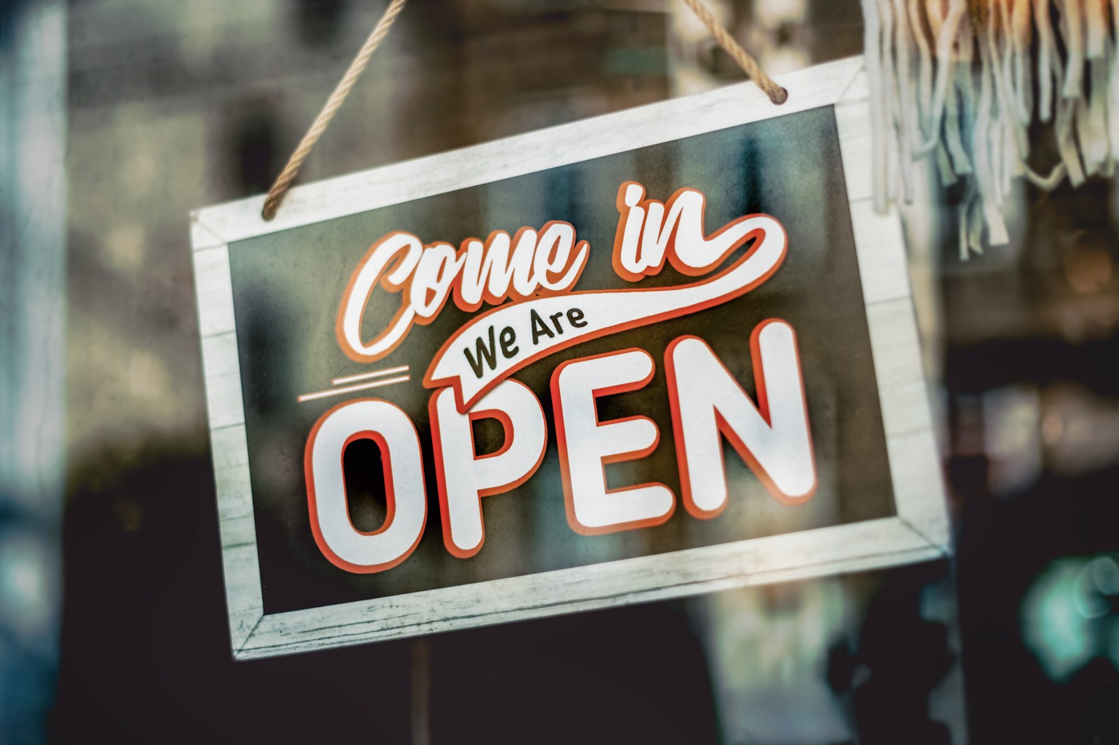 A sign hanging in a shop window reads Come in, we are open, against a blurred interior