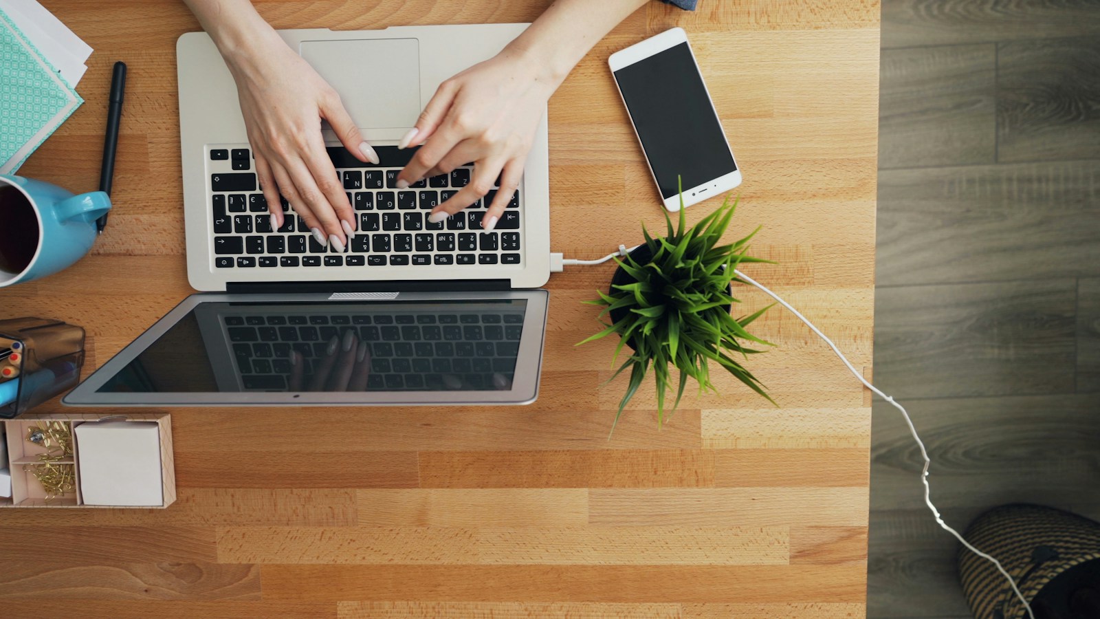 Hands type on a laptop on a wooden desk beside a second laptop, a white smartphone and a potted plant
