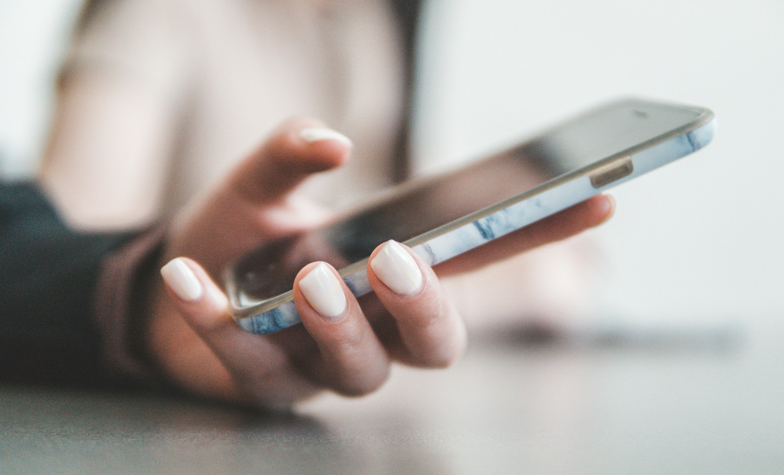 Close-up of a hand with white painted nails holding a smartphone in a marble case, ready to tap the screen