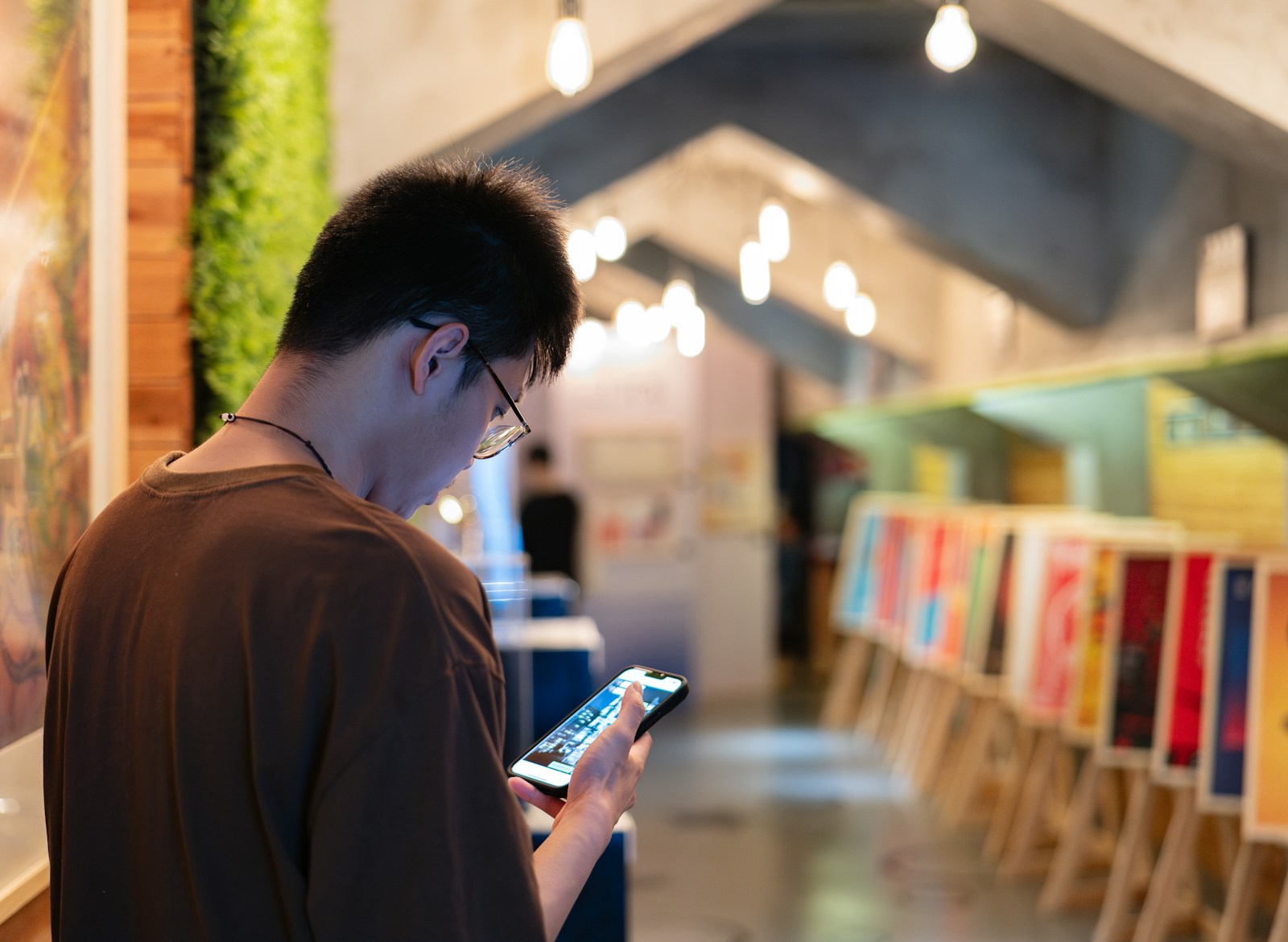 A young man in glasses reads his smartphone while standing in a shop lined with framed posters