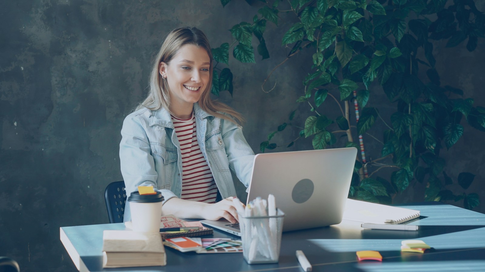 A smiling woman in a denim jacket types on a laptop at a desk with a coffee cup, notebooks and a pot of pens