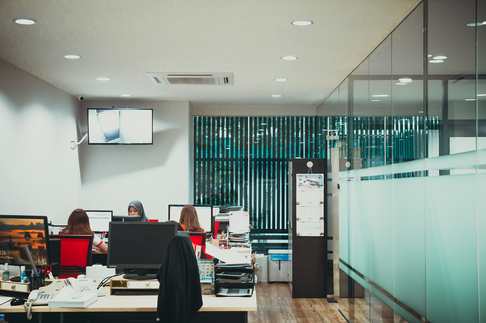 Several women work at desktop computers in a small office, two corded desk phones in the foreground