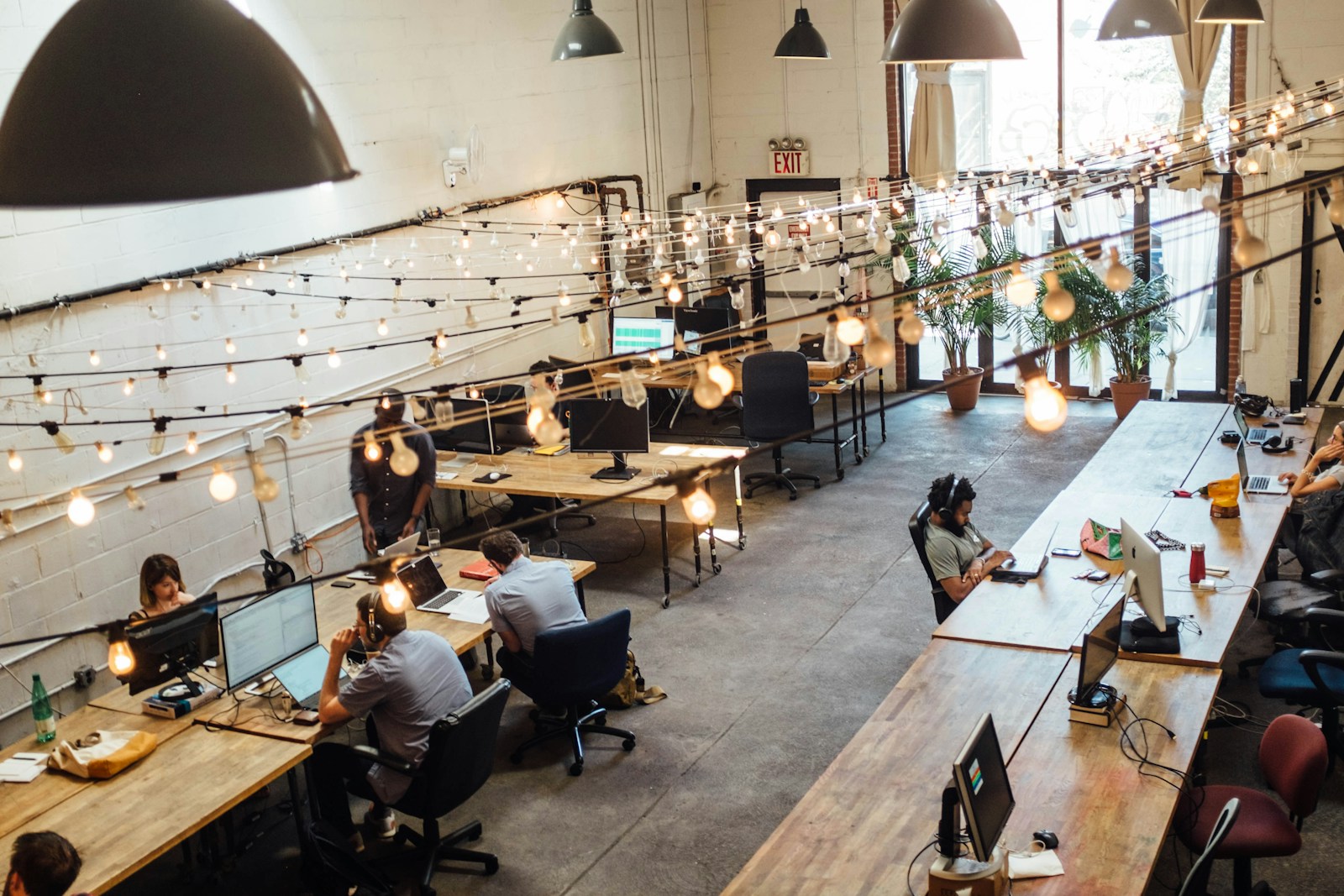 String lights hang over long wooden desks in a converted warehouse office where several people work at laptops and monitors