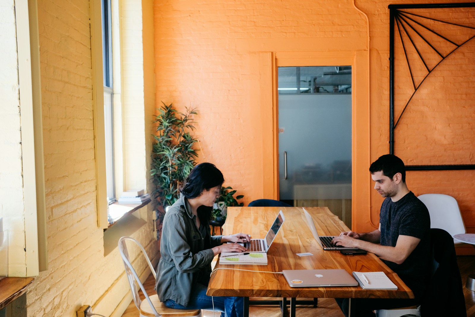 A woman and a man type on separate laptops at a long wooden table in an orange-walled office