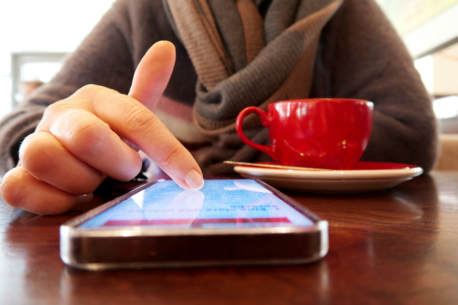 A finger taps a smartphone lying on a café table next to a red espresso cup and saucer