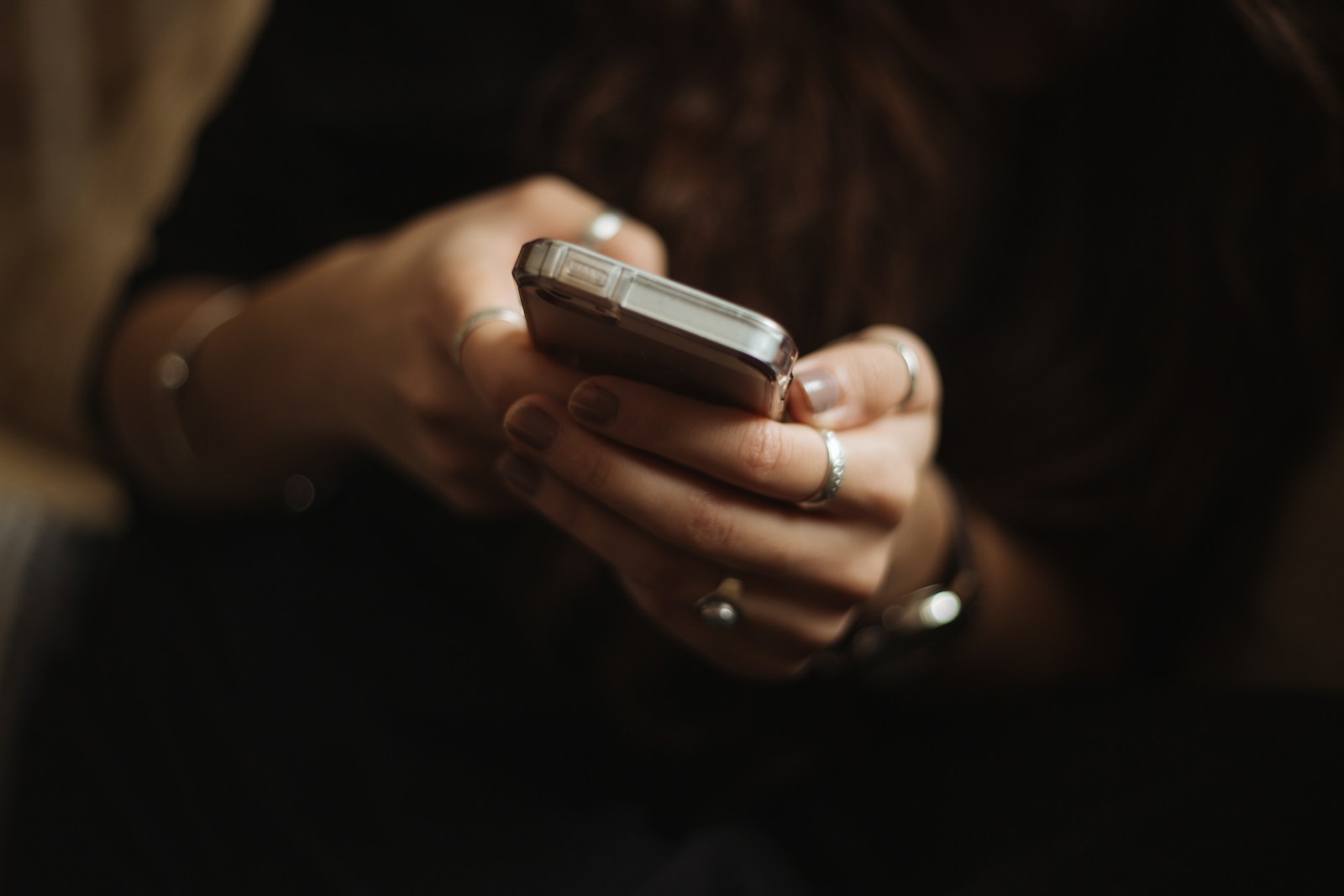Close-up of a person's hands, wearing silver rings, holding a smartphone and typing in a dimly lit room