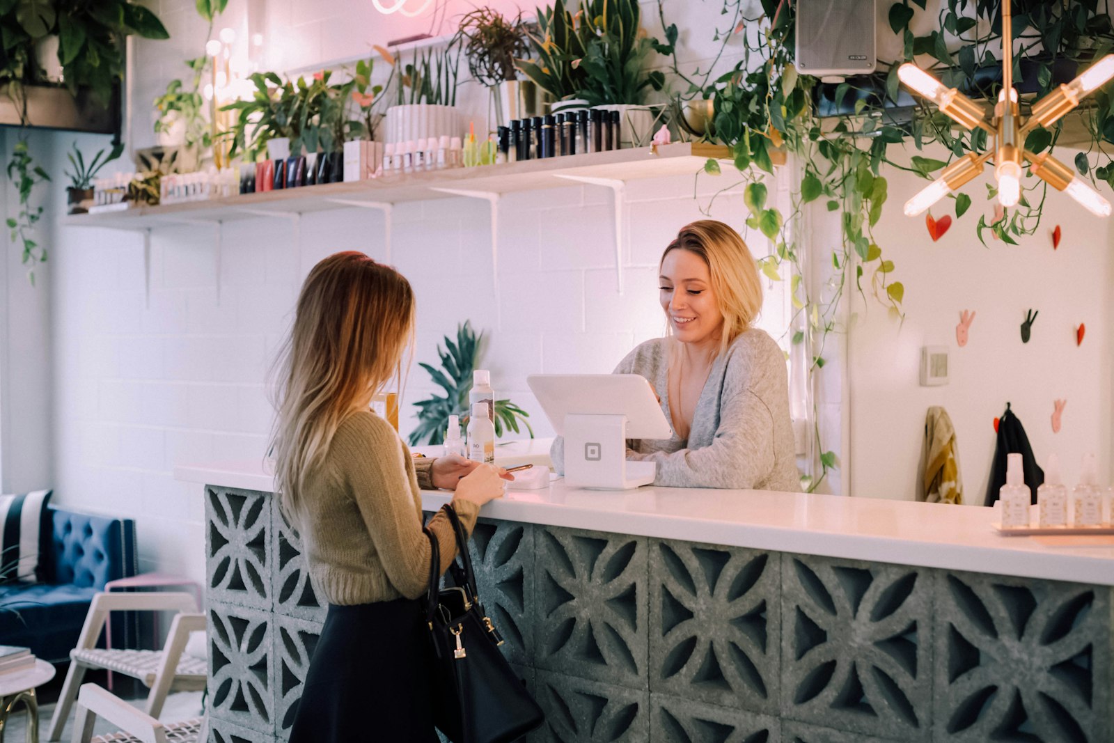 A customer at a white shop counter talks with a smiling assistant behind a card reader in a plant-filled store