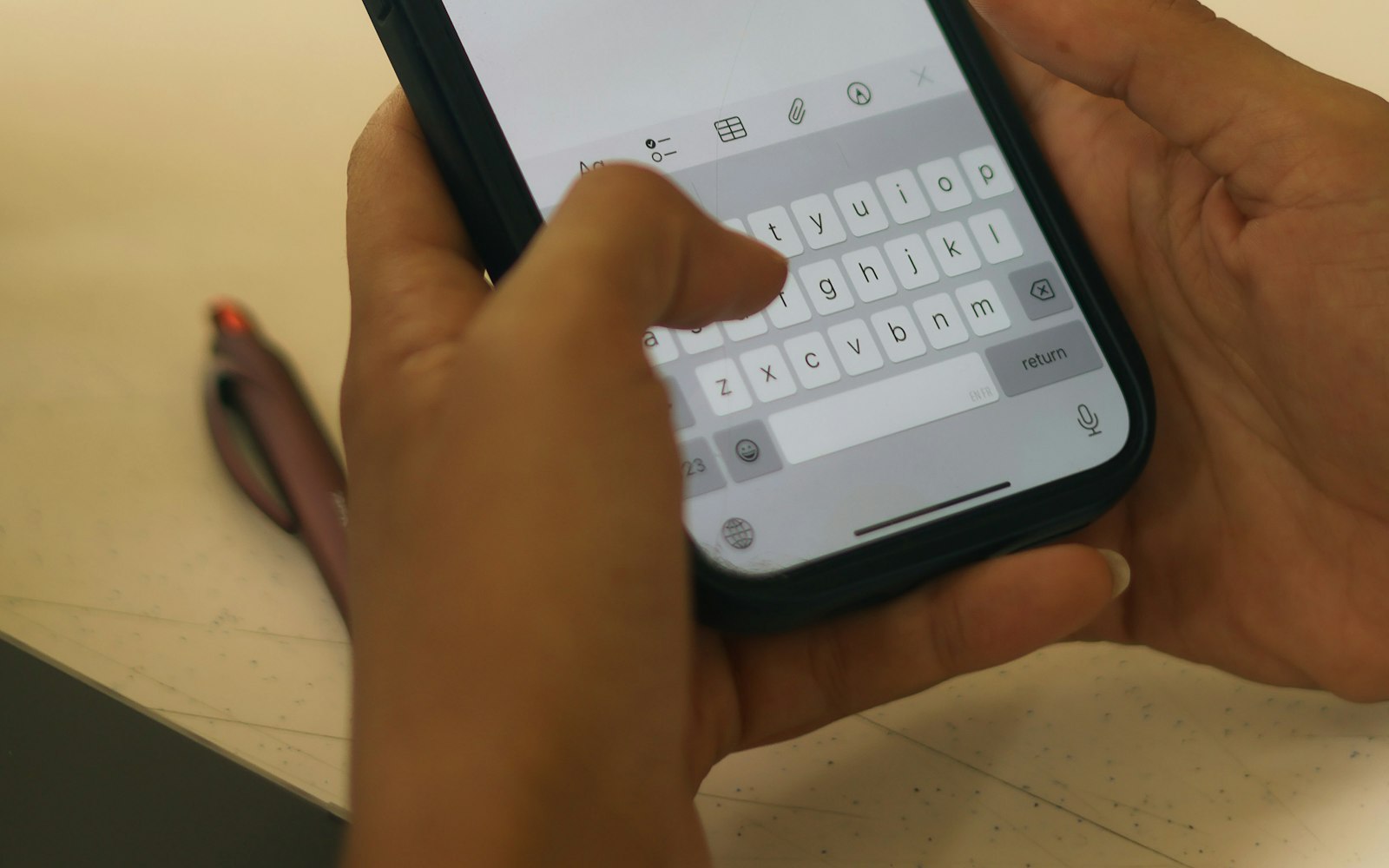 Close-up of two hands typing a message on a smartphone keyboard, with a pen lying on the desk beside them