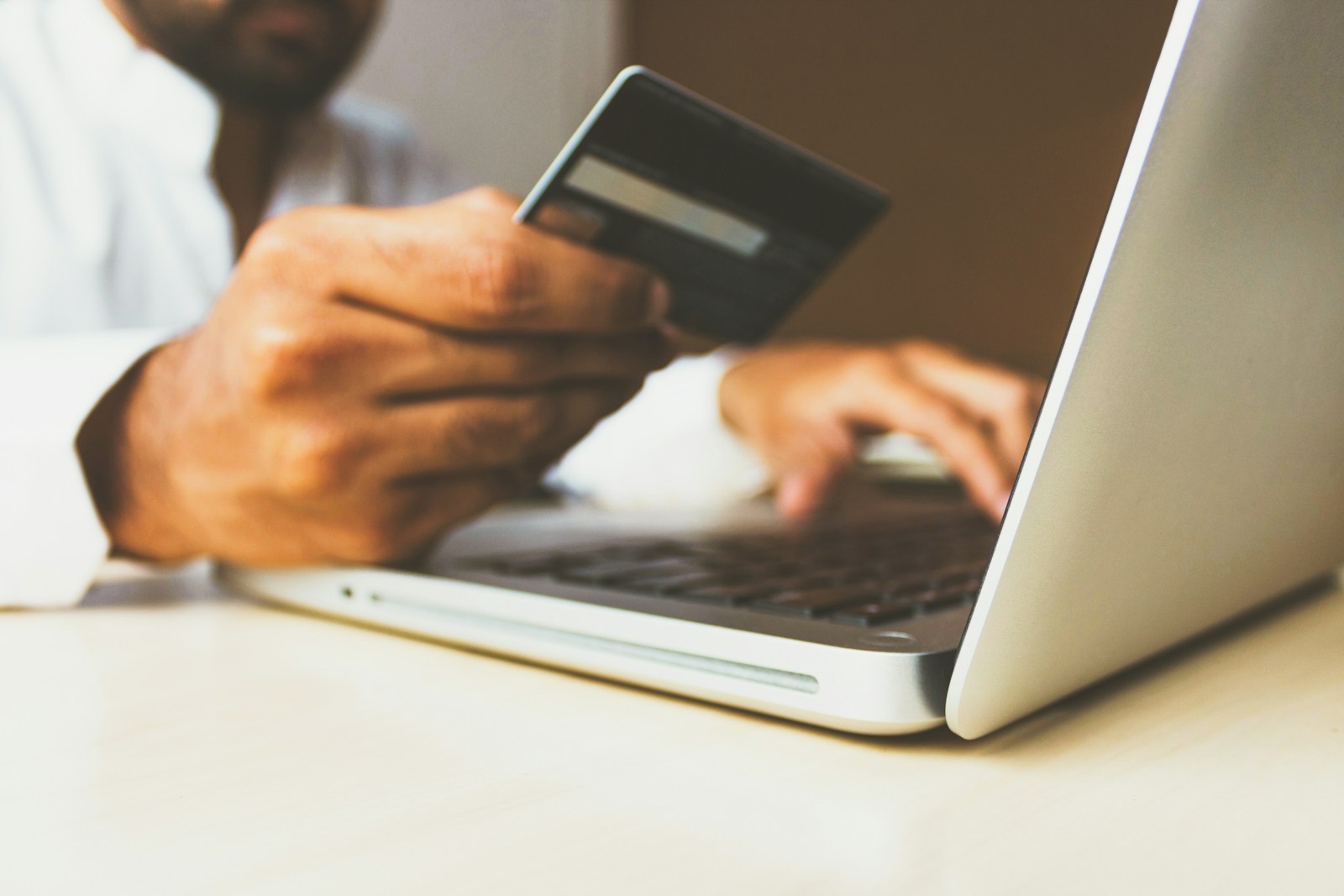 A man in a white shirt holds a bank card in one hand while typing on a silver laptop at a light wooden desk