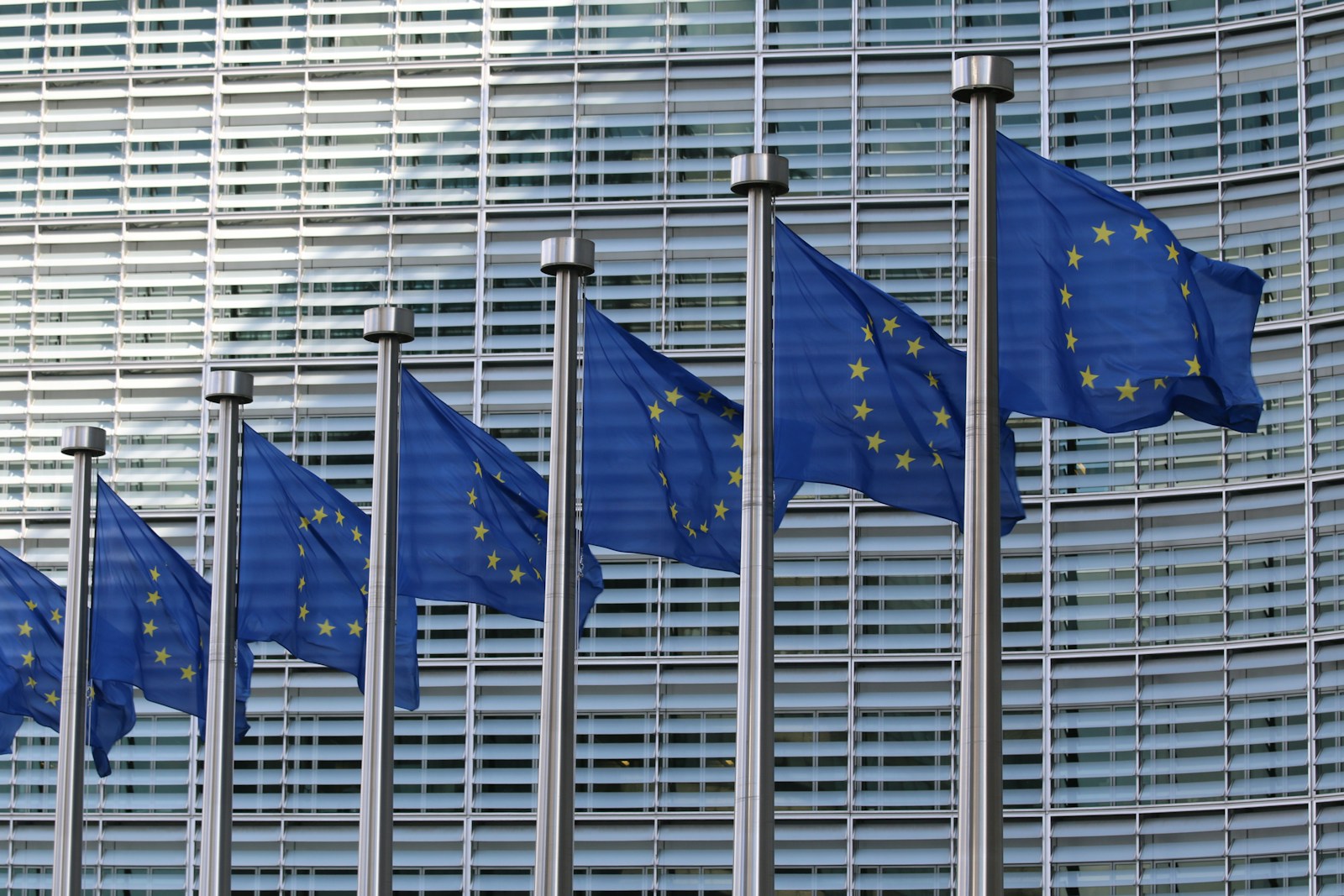 A row of blue European Union flags with gold stars on steel poles in front of a curved glass office facade