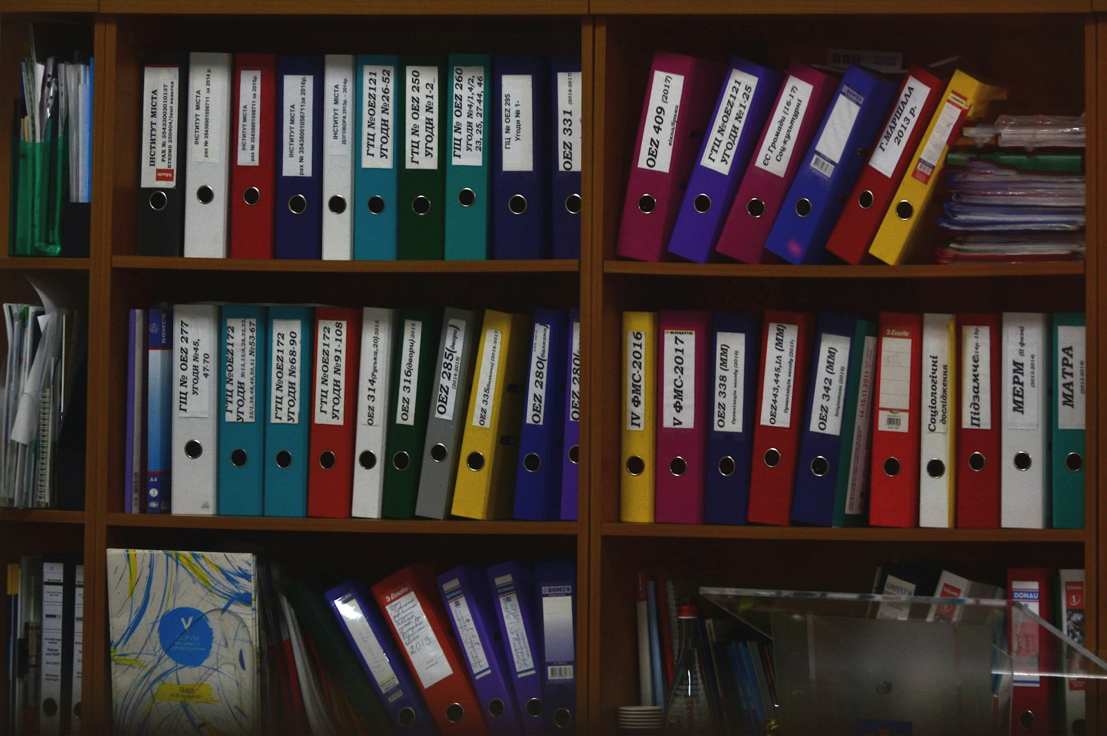 Rows of colourful ring binders with labelled spines fill the shelves of a wooden office bookcase
