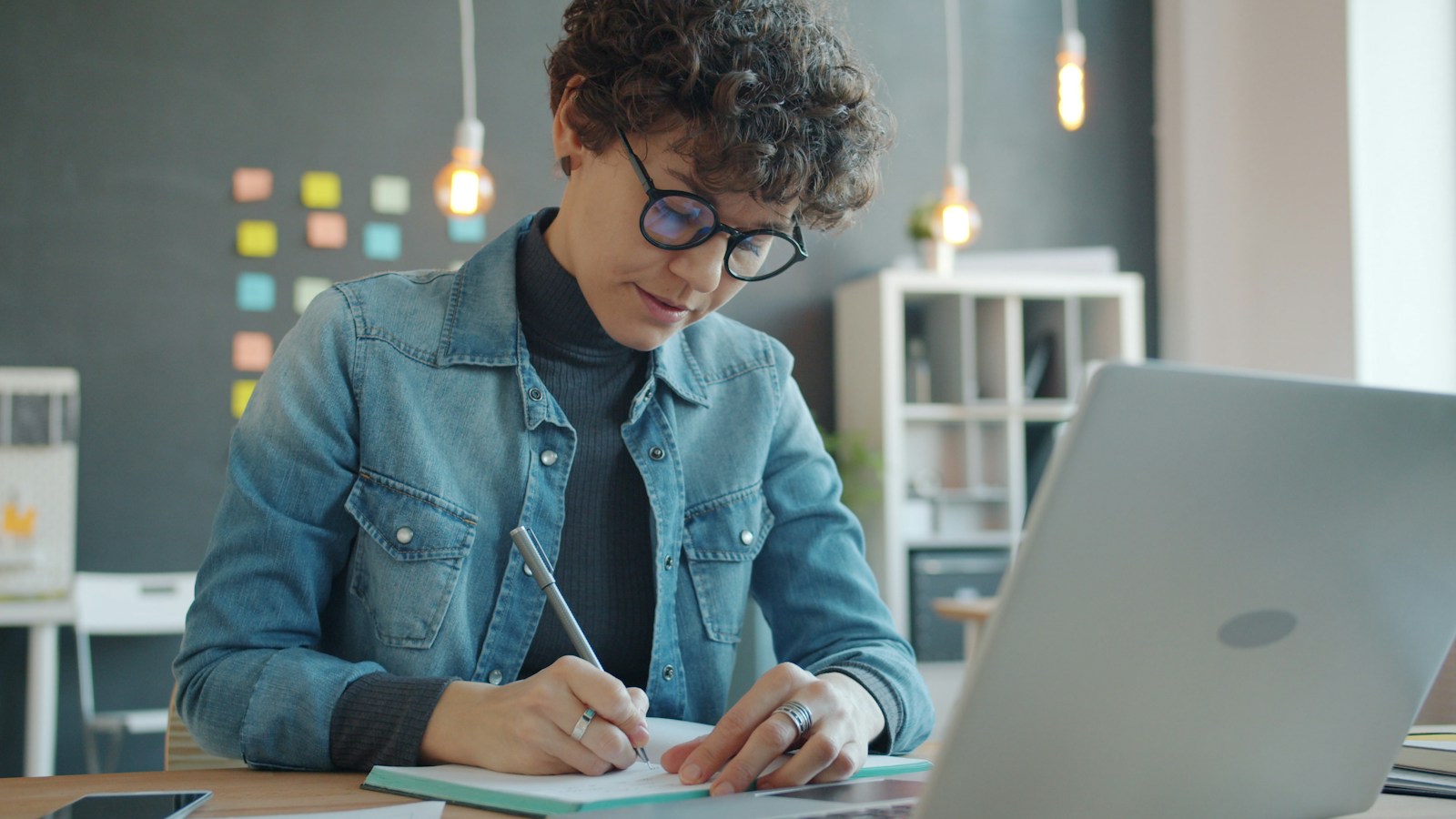 A woman in glasses writes in a notebook beside an open laptop, with sticky notes on the wall behind