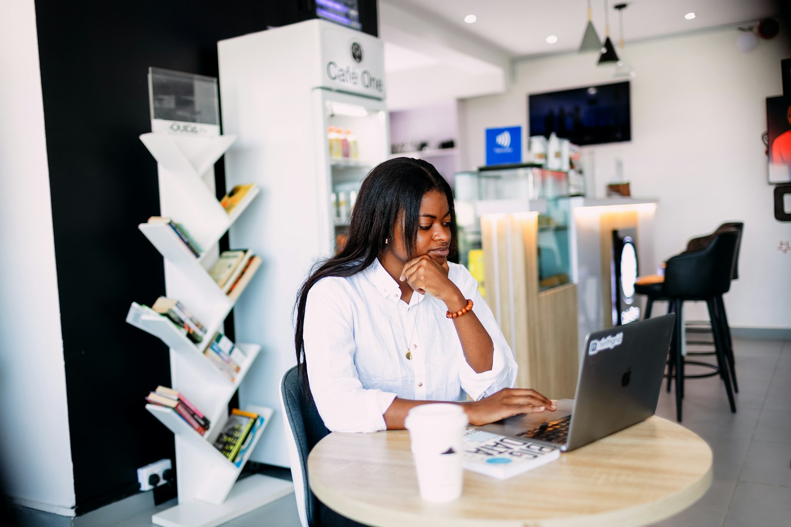 A woman in a white shirt works on a laptop at a round café table next to a takeaway coffee cup
