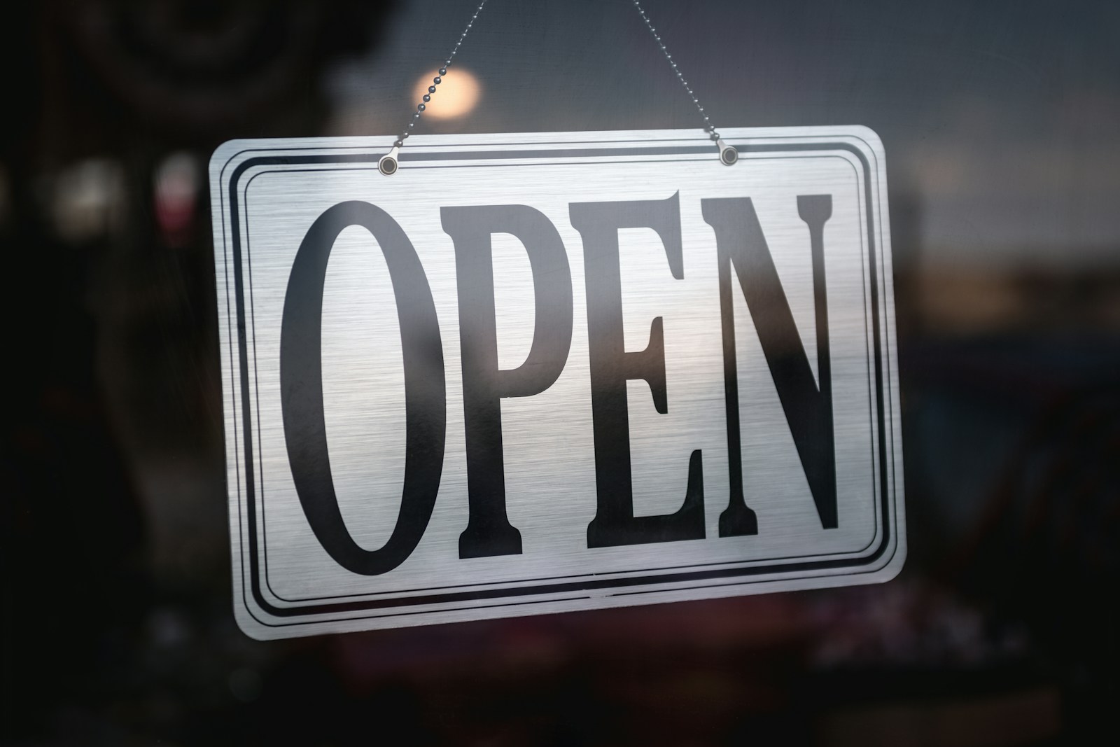 A silver OPEN sign hangs on a chain inside the glass door of a dimly lit shop