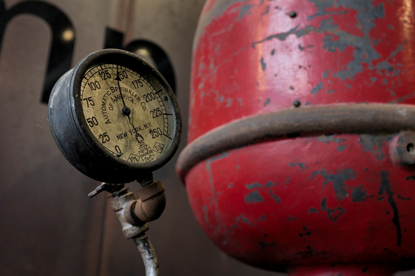 A weathered round pressure gauge with a black needle mounted on a pipe beside a scuffed red metal tank
